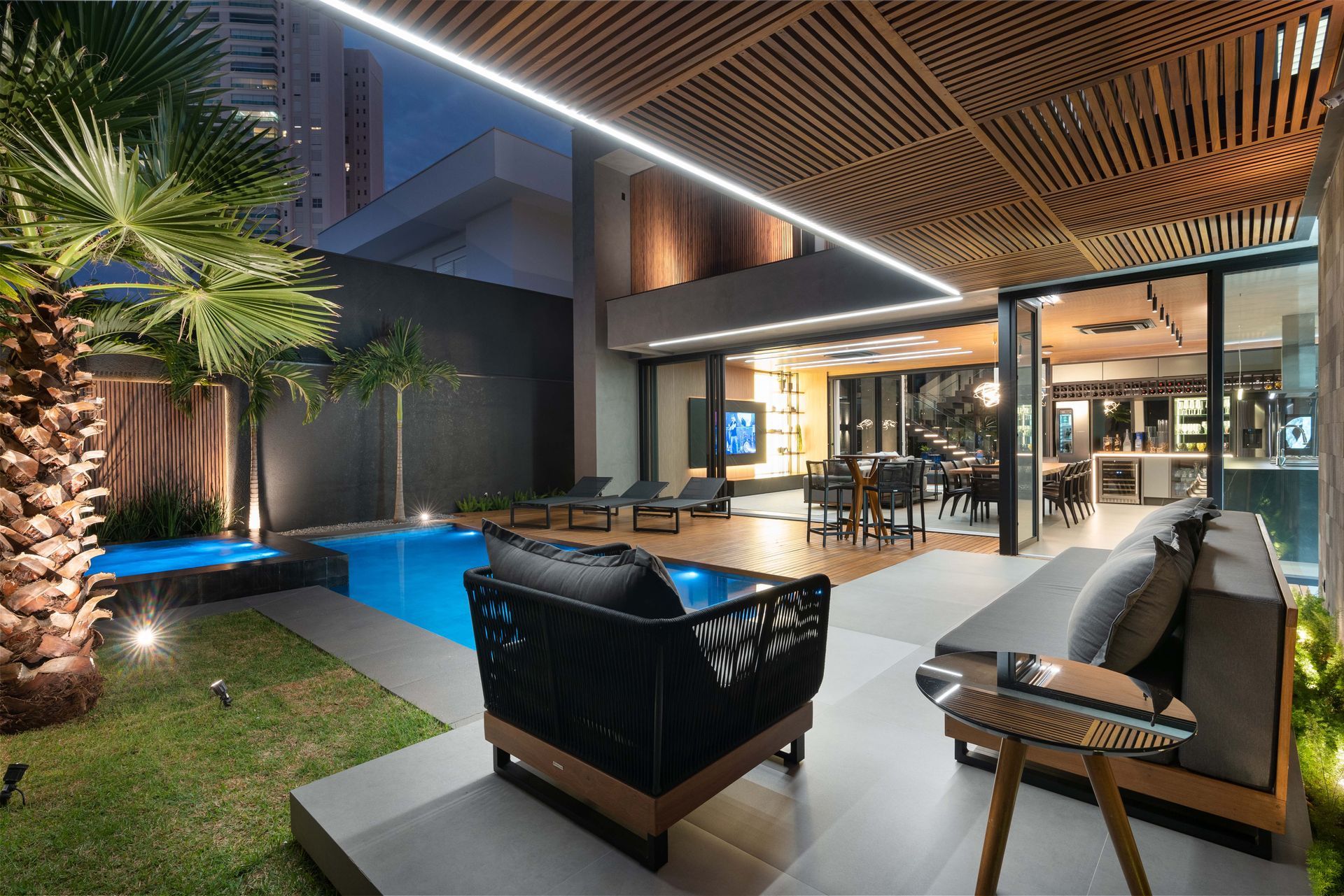 Pátio externo com piscina, área de estar e sala de jantar. Design moderno com detalhes em madeira e tons escuros à noite.