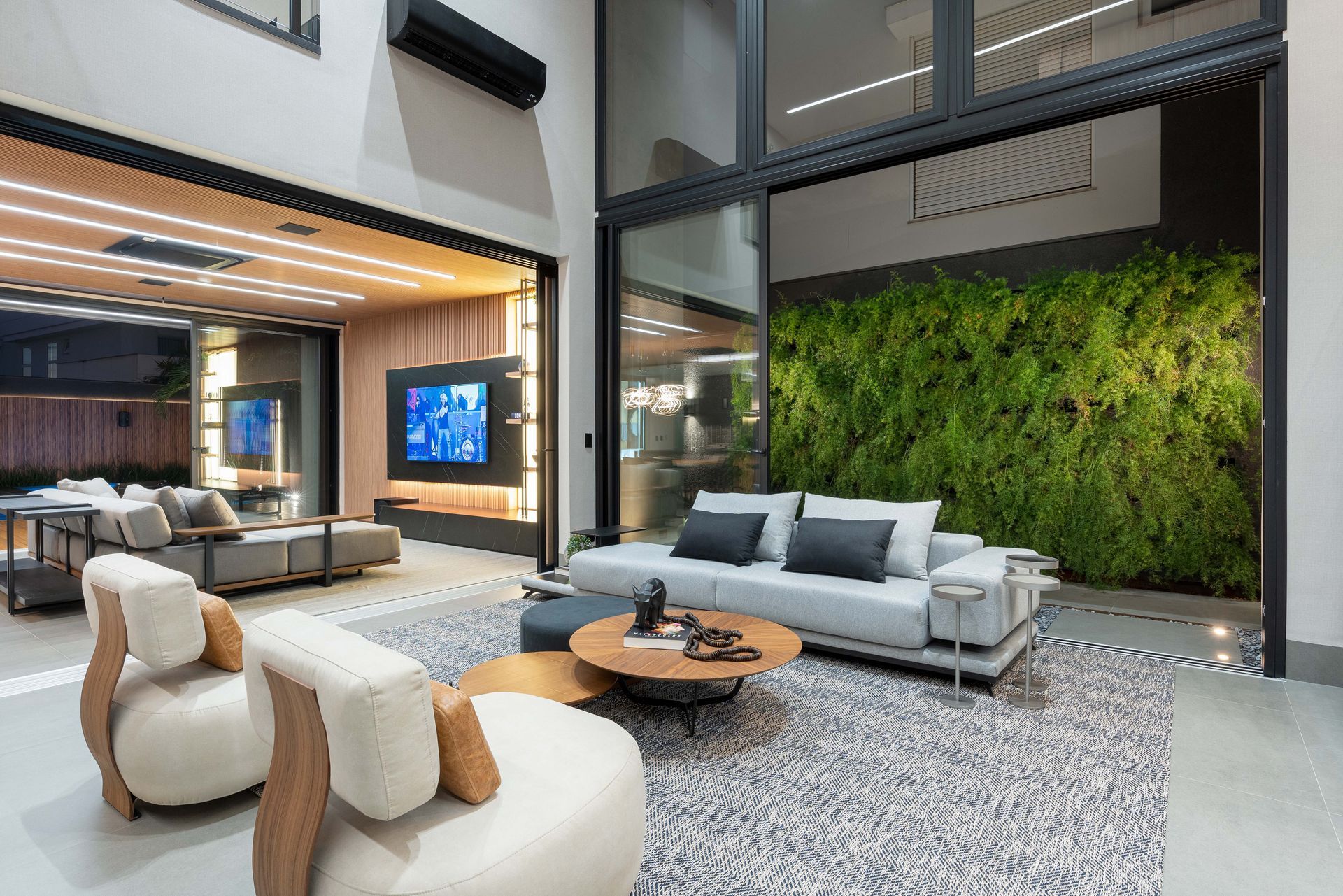 Sala de estar luxuosa com portas abertas para uma parede verde. Possui um sofá cinza, mesa de centro redonda e poltronas sobre um tapete estampado.