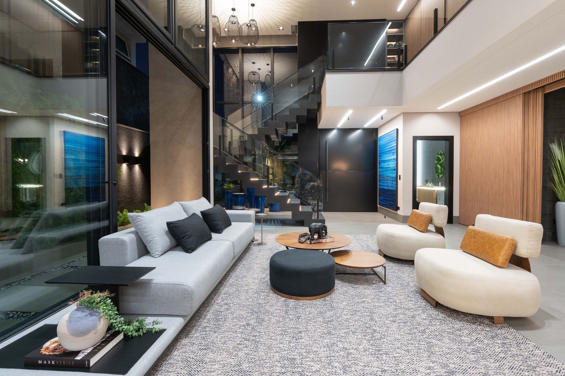 Sala de estar moderna com sofá cinza, cadeiras brancas, tapete e uma escada de vidro.