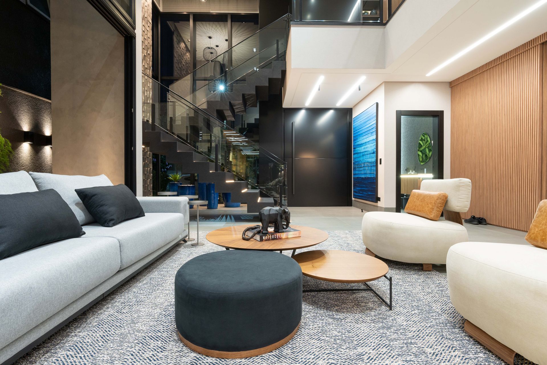 Sala de estar moderna com sofá cinza, cadeiras brancas, mesa de centro, escada e grande quadro.