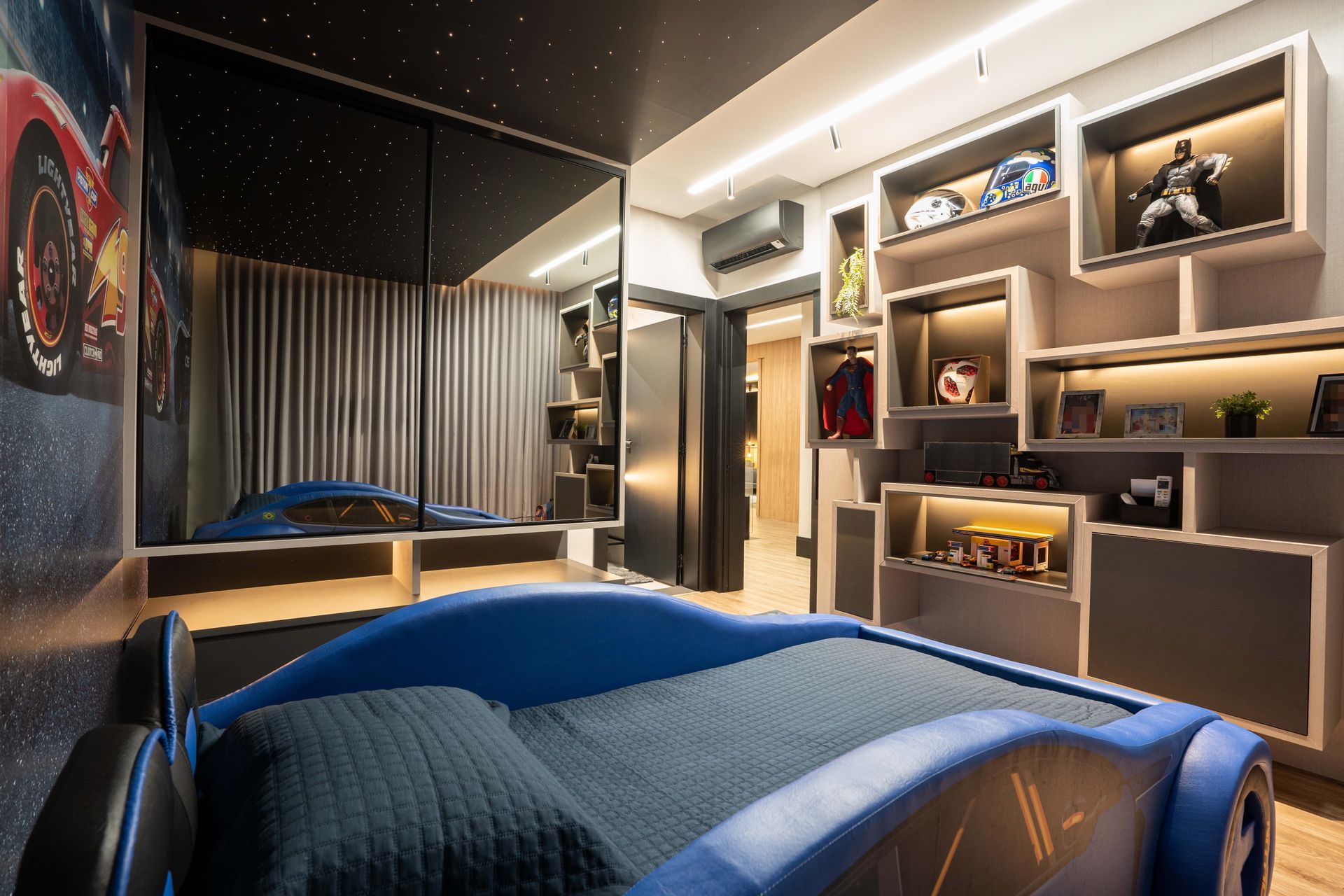 Uma cama azul com tema de carro de corrida em um quarto temático com prateleiras de exposição e um mural de carros.