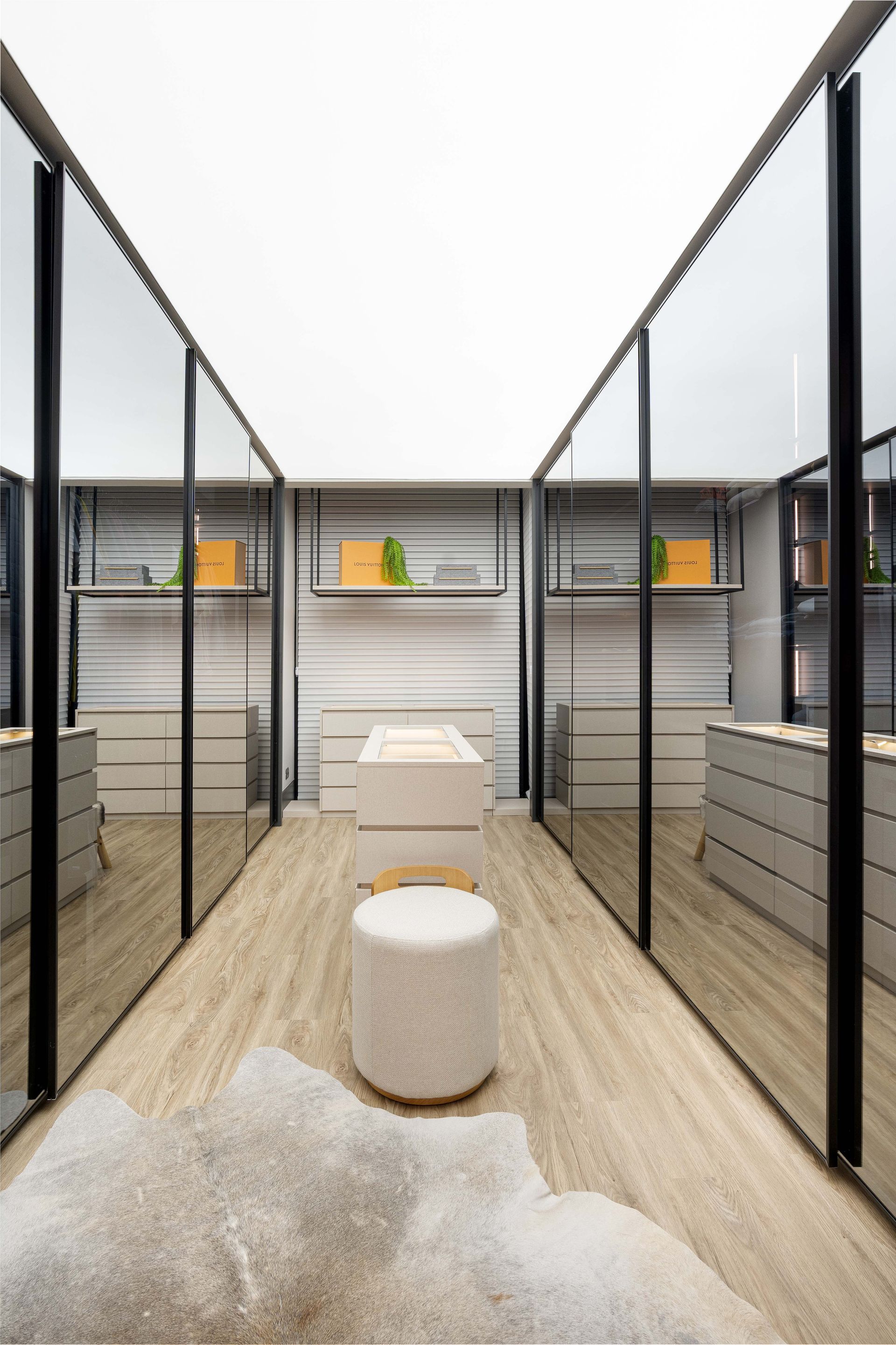 Uma sala longa e estreita com paredes espelhadas, piso de madeira de cor clara e um tapete decorativo.