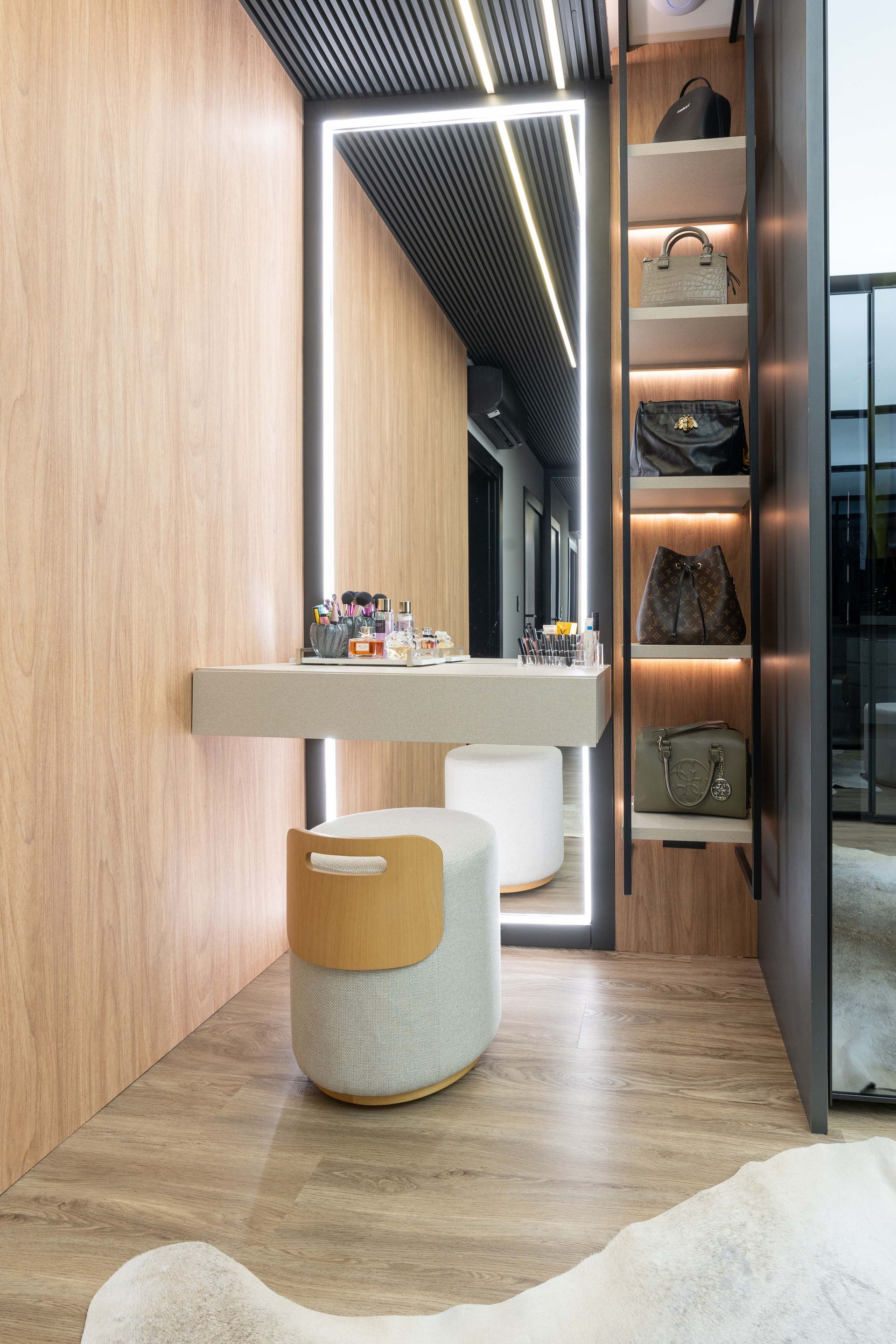 Área de penteadeira com painéis de madeira, espelho iluminado, banco e prateleiras para bolsas.