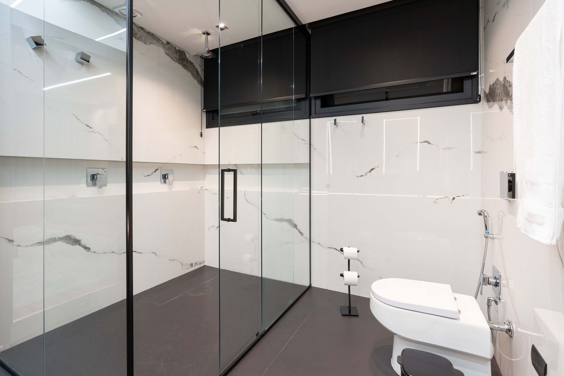 Banheiro moderno com box de vidro, paredes de mármore branco, detalhes em preto e vaso sanitário.