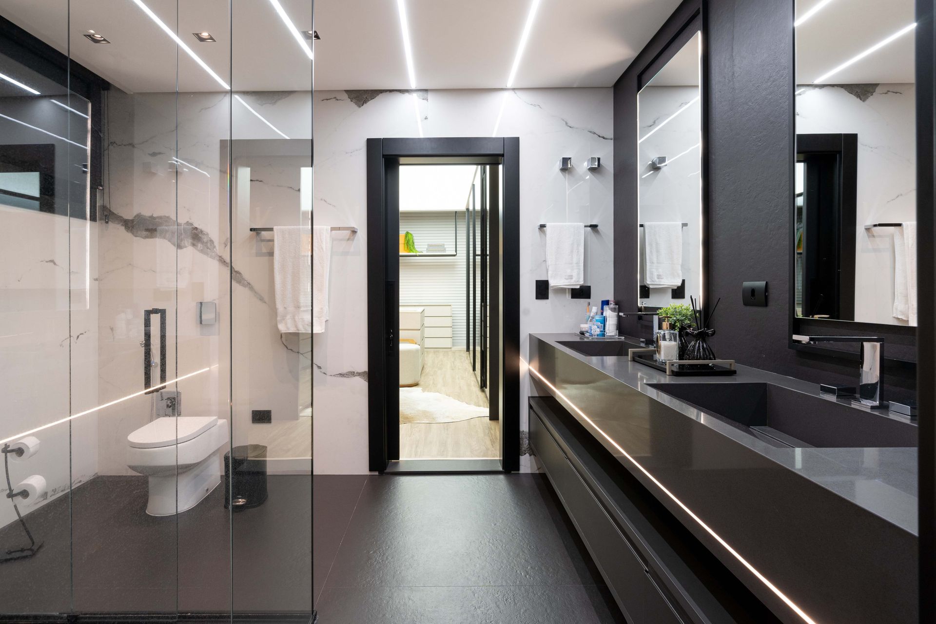 Banheiro moderno com elementos em preto e cinza. Possui box de vidro, pias duplas e iluminação linear.