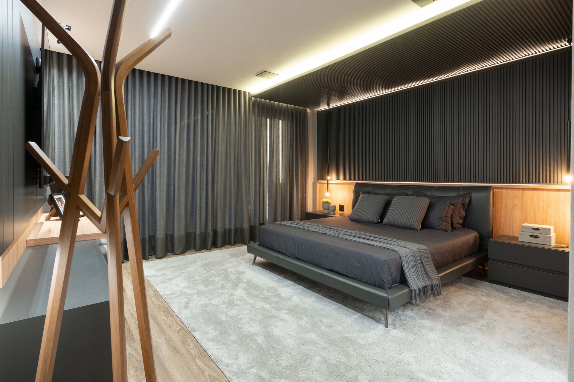 Quarto escuro com cama cinza, cortinas e carpete. Cabideiro de madeira.
