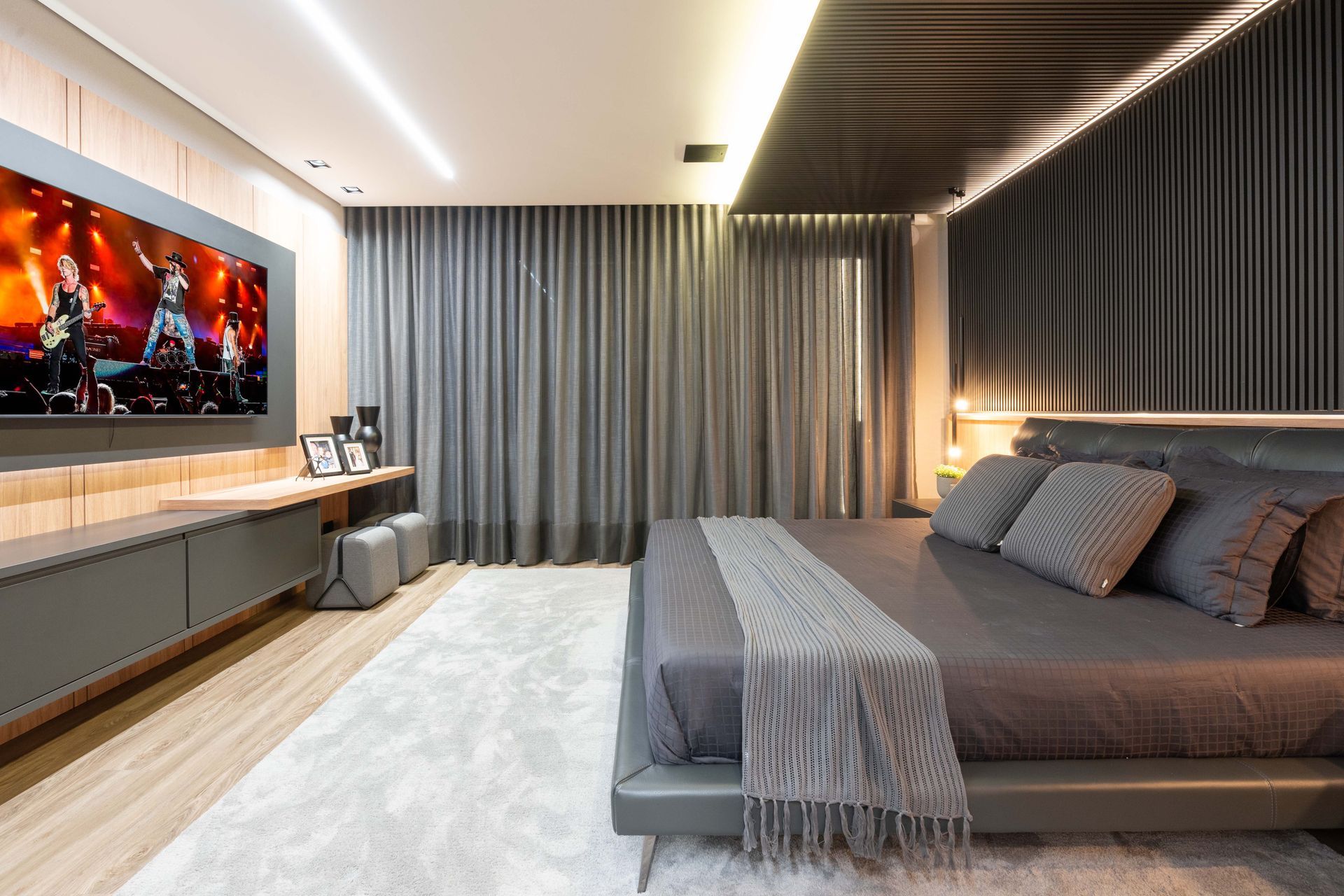 Quarto cinza moderno com TV grande, cama e cortinas.