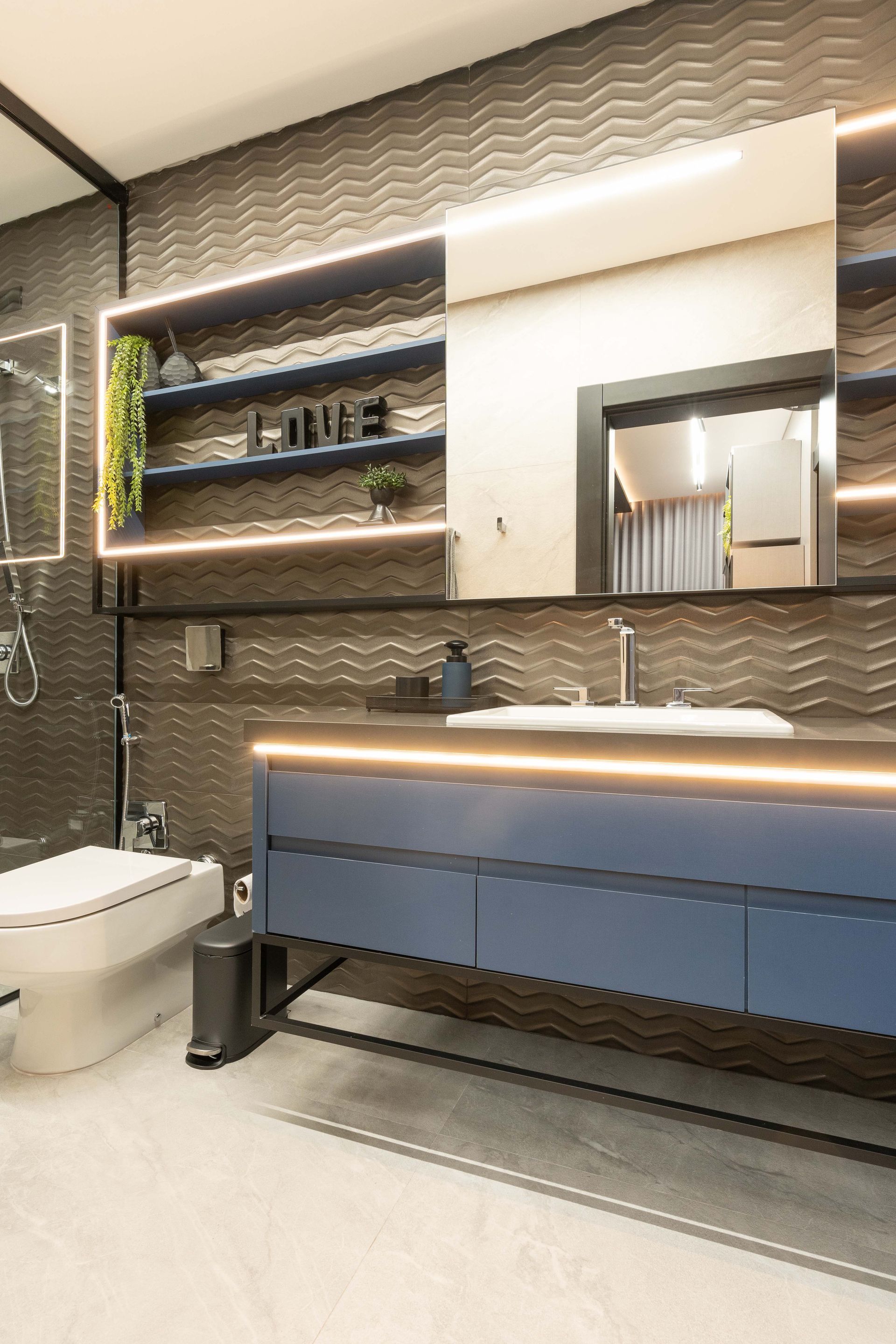 Banheiro moderno com penteadeira azul, parede escura texturizada, espelho, prateleiras com plantas e vaso sanitário.