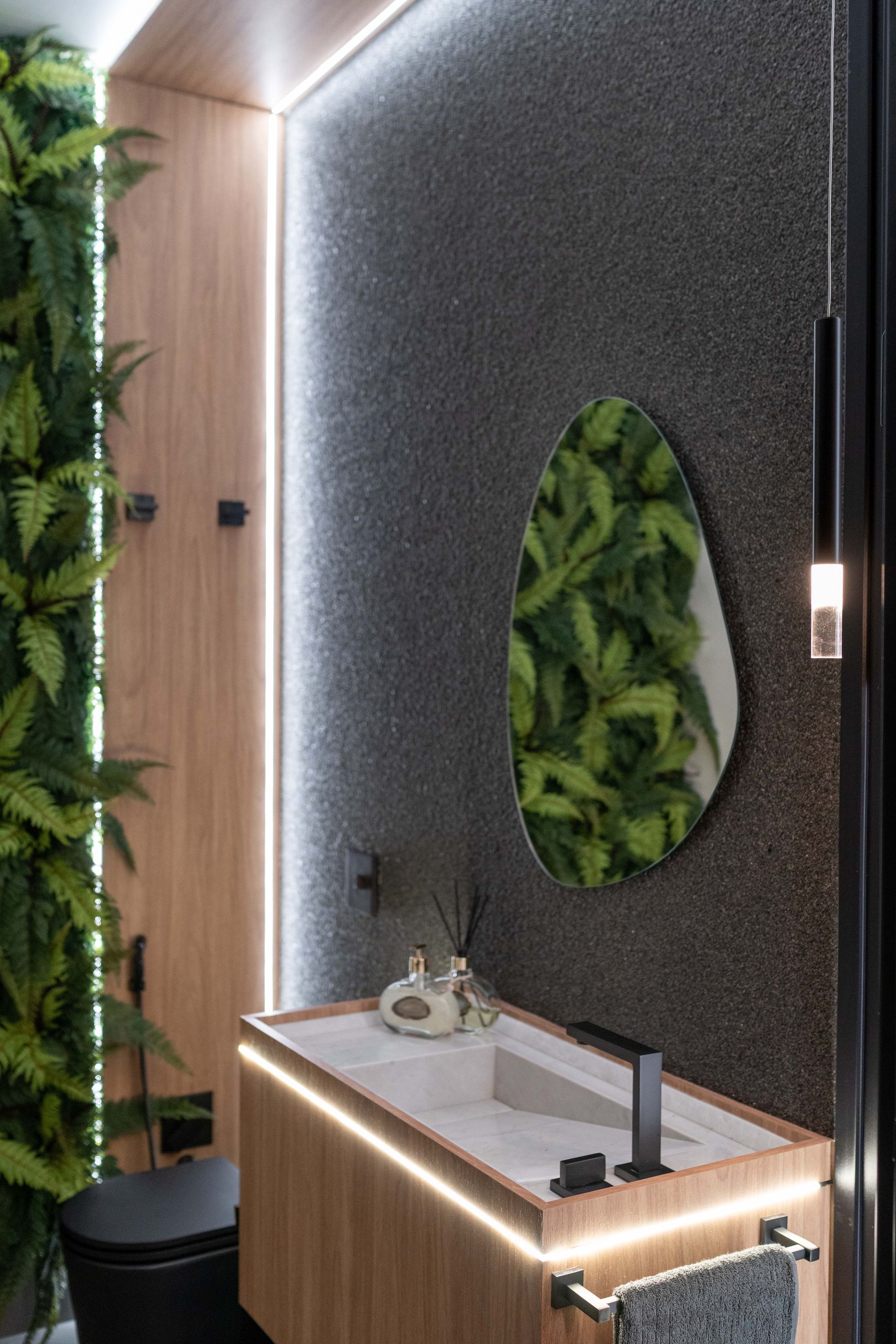 Banheiro moderno com paredes de madeira e textura escura, iluminado por luzes de LED. Possui espelho e pia com plantas.