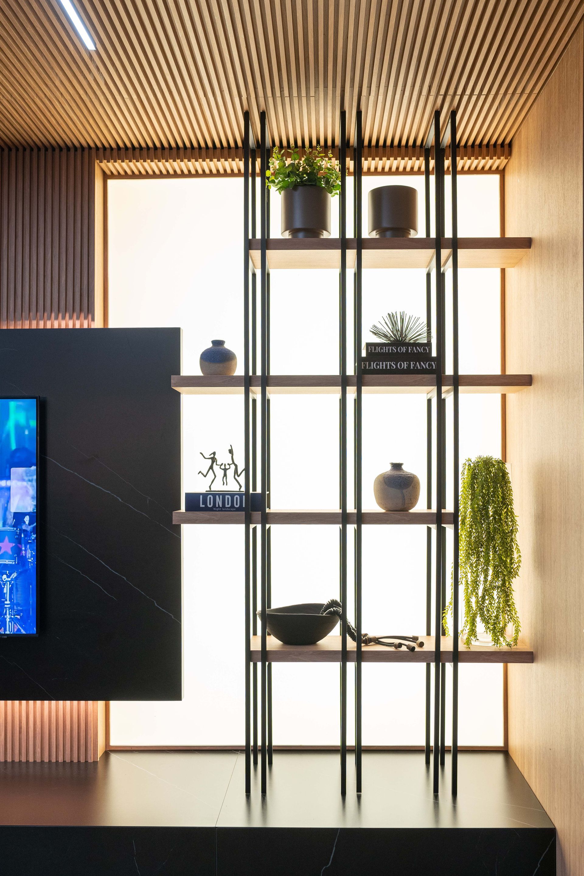 Estante com plantas e decoração em painel claro; estrutura escura, prateleiras de madeira, tons neutros.