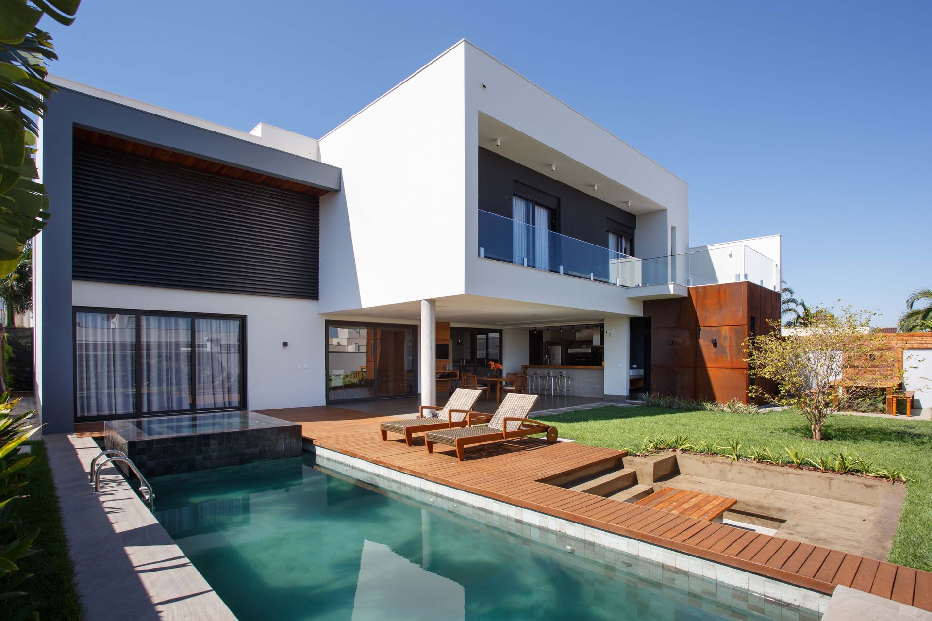 Casa branca moderna com piscina e deck de madeira, duas espreguiçadeiras, gramado verde, dia ensolarado.