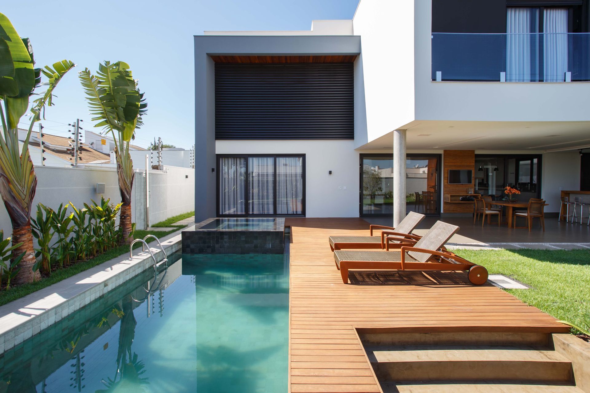 Casa moderna com piscina, deck de madeira e espreguiçadeiras; ambiente externo ensolarado.