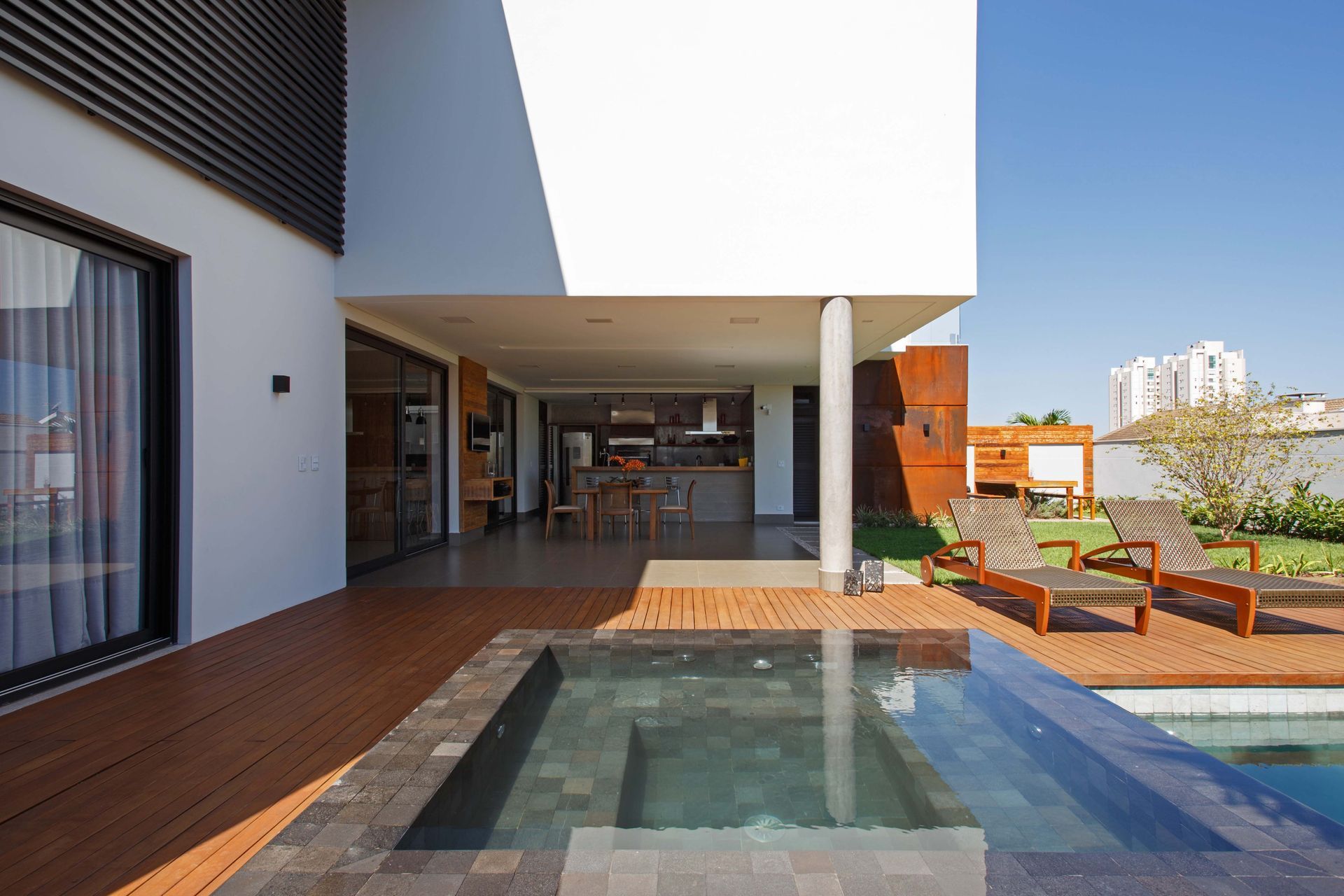 Casa moderna com piscina, deck de madeira e cozinha ao ar livre, sob um céu azul.