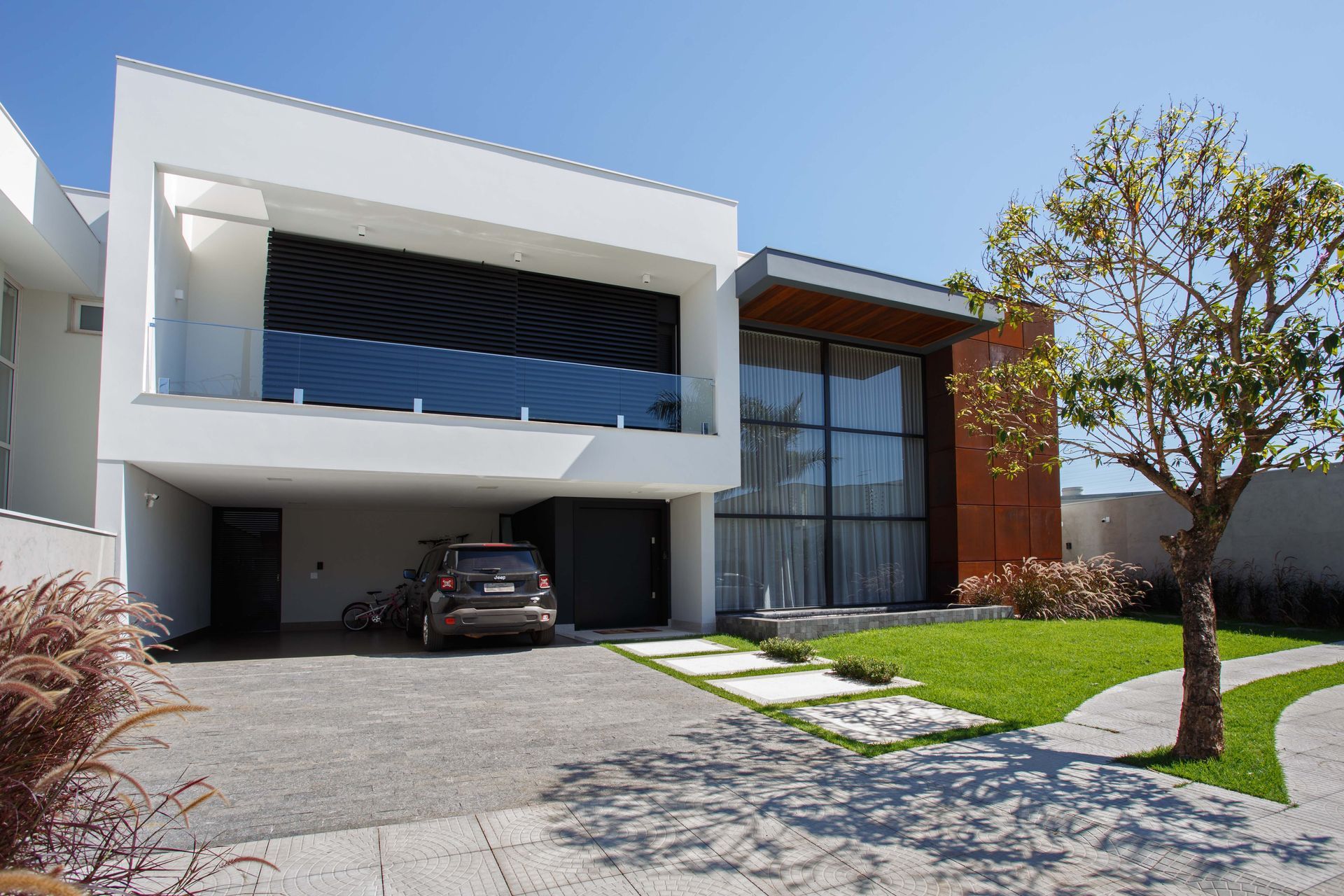 Casa branca moderna com um carro estacionado na garagem e uma pequena árvore na frente.