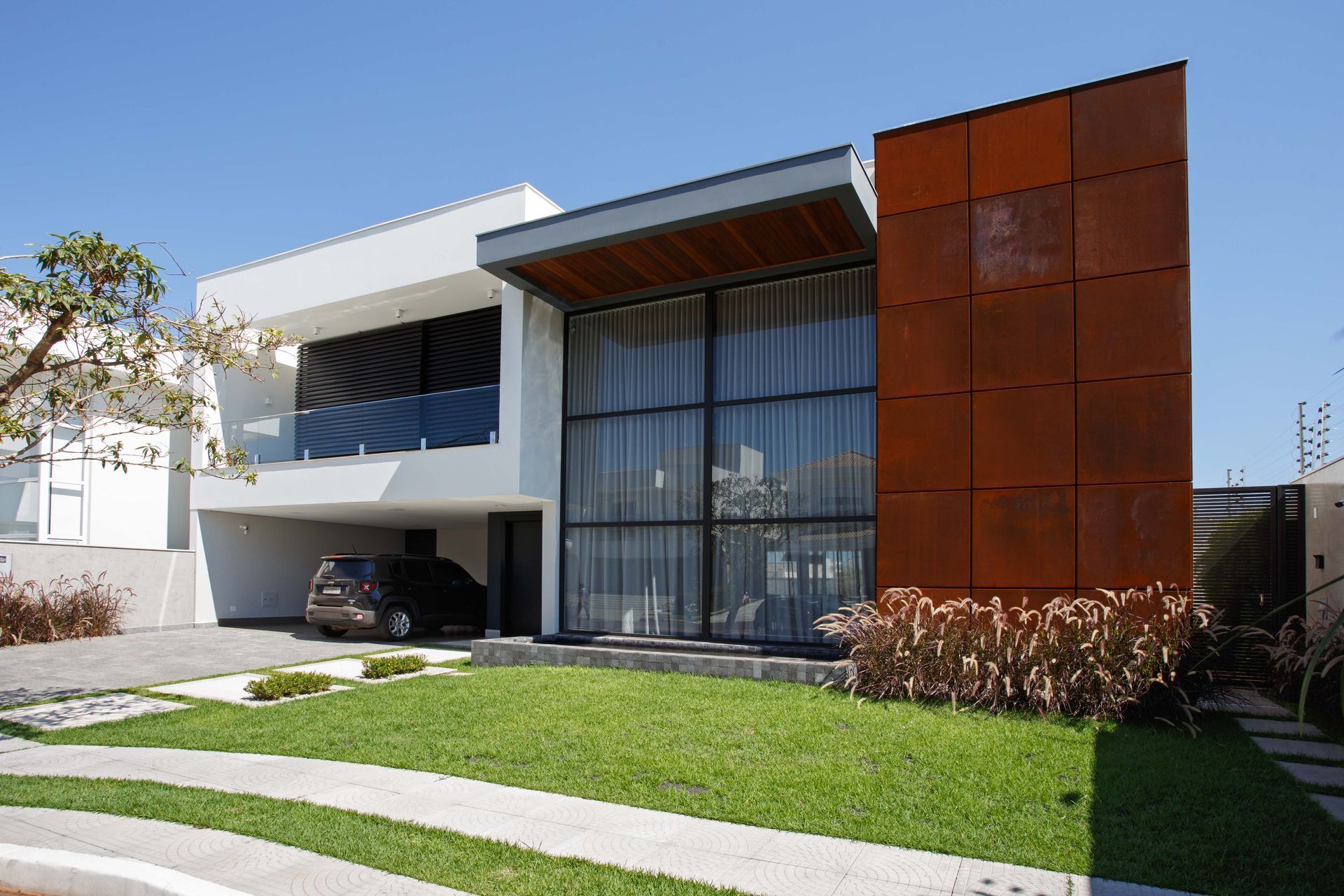 Casa branca moderna com fachada de metal enferrujada, frente de vidro e carro estacionado em uma garagem coberta em um dia ensolarado.