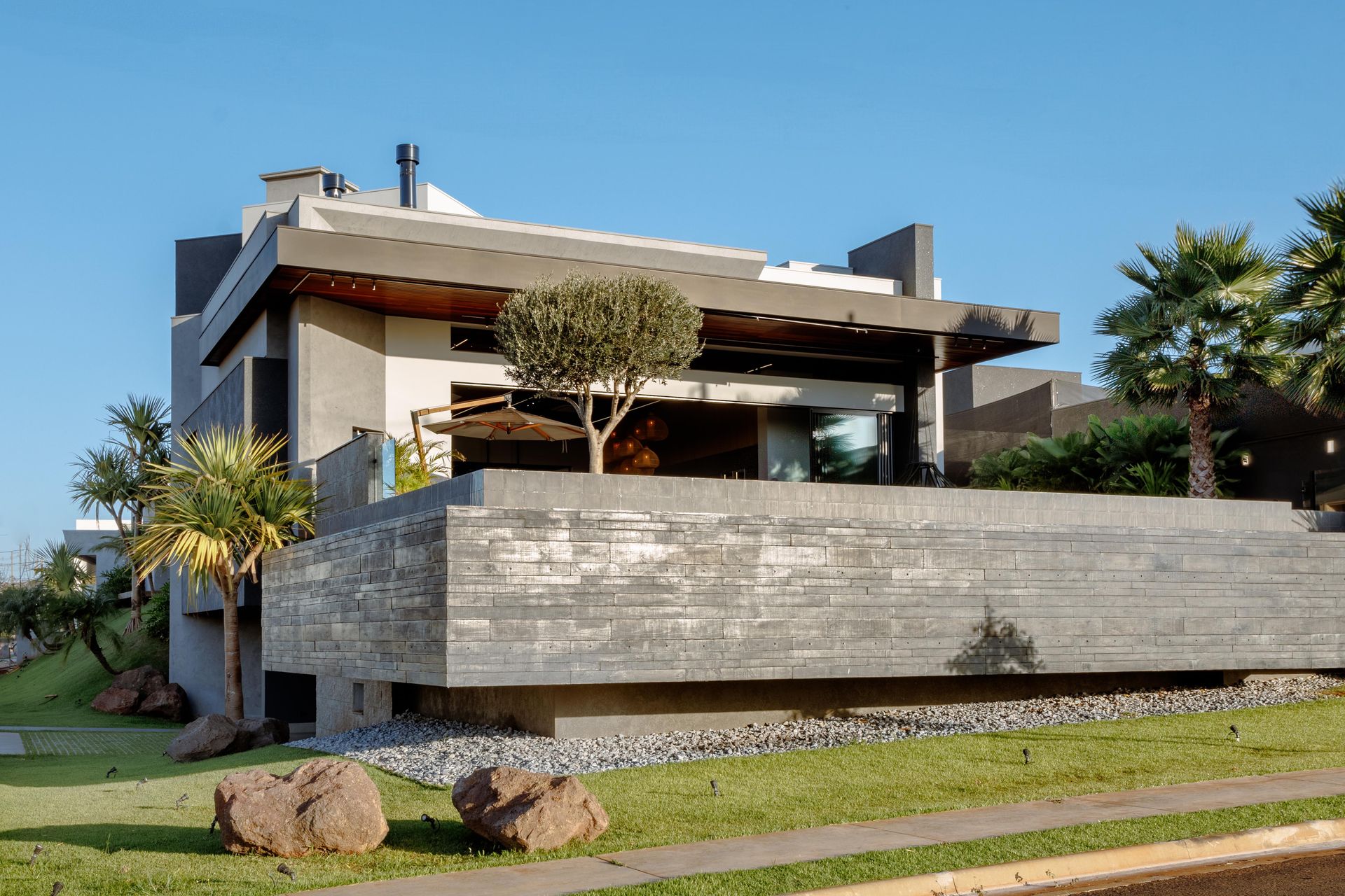 Casa moderna de concreto com telhado plano, em um dia ensolarado. Palmeiras e um gramado na frente.