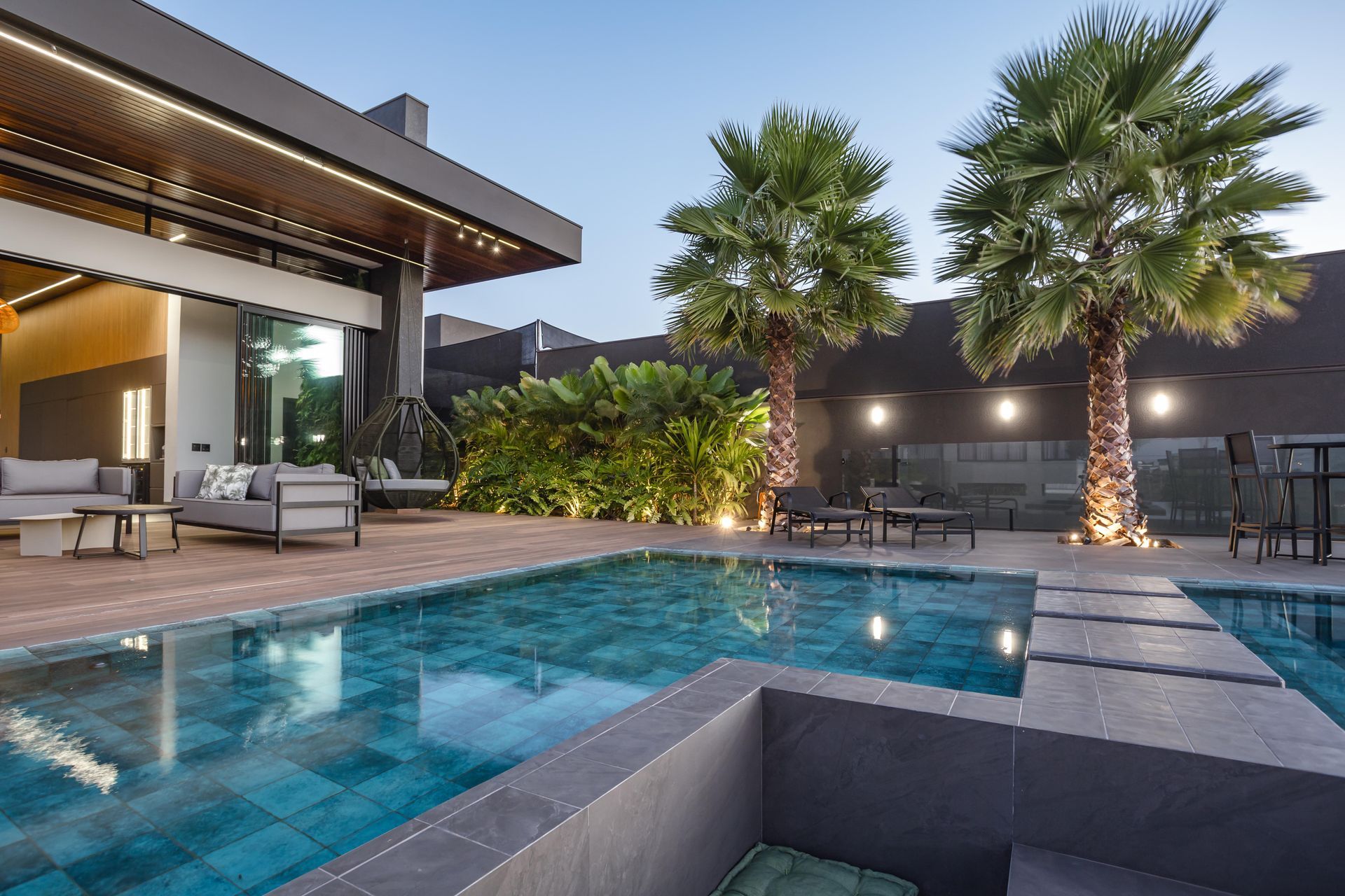 Área de estar à beira da piscina ao entardecer com palmeiras, sofás e piscina.