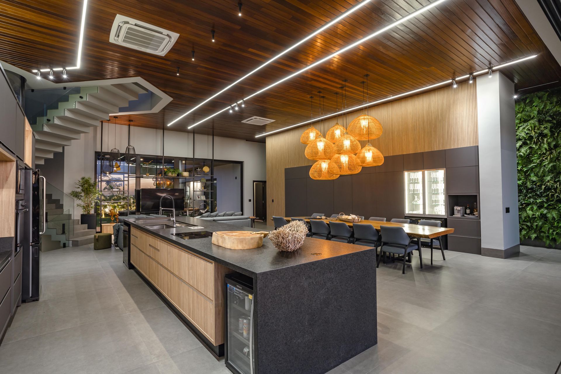 Cozinha moderna com ilha grande, armários de madeira e área de jantar. Escada para o andar superior e parede verde.
