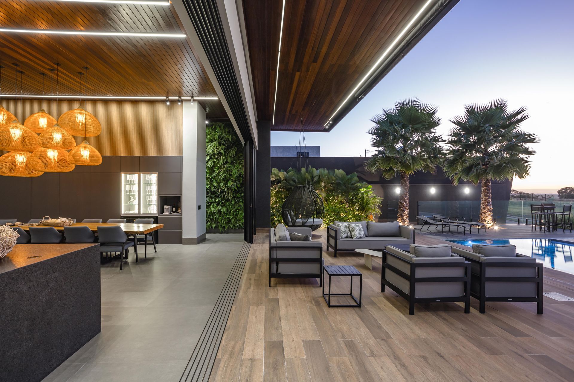 Pátio externo com mesas à beira da piscina. Teto de madeira, parede verde e palmeiras.