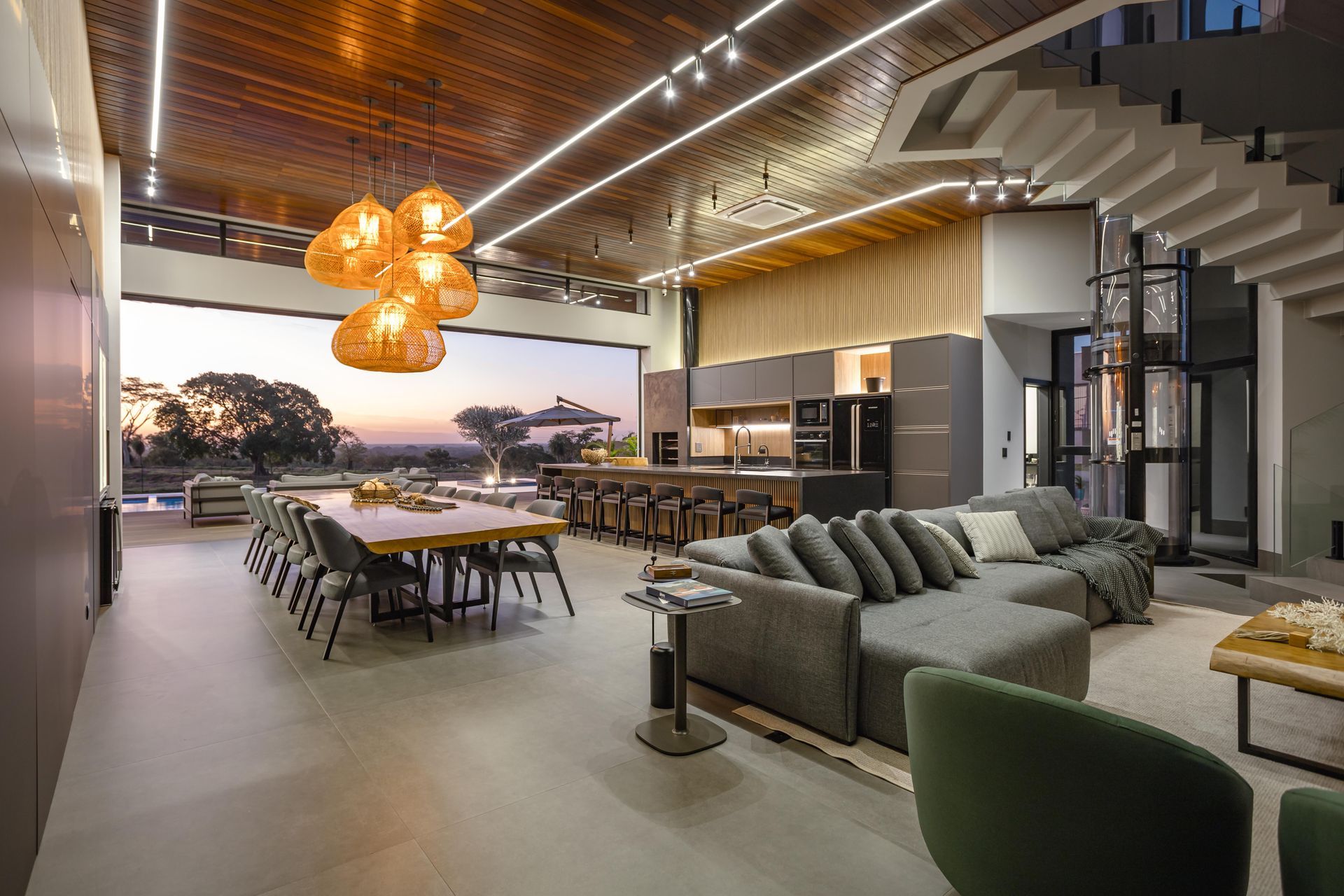 Interior moderno e aberto com salas de jantar, cozinha e estar. Amplas janelas com vista para o exterior, teto de madeira e cores neutras.