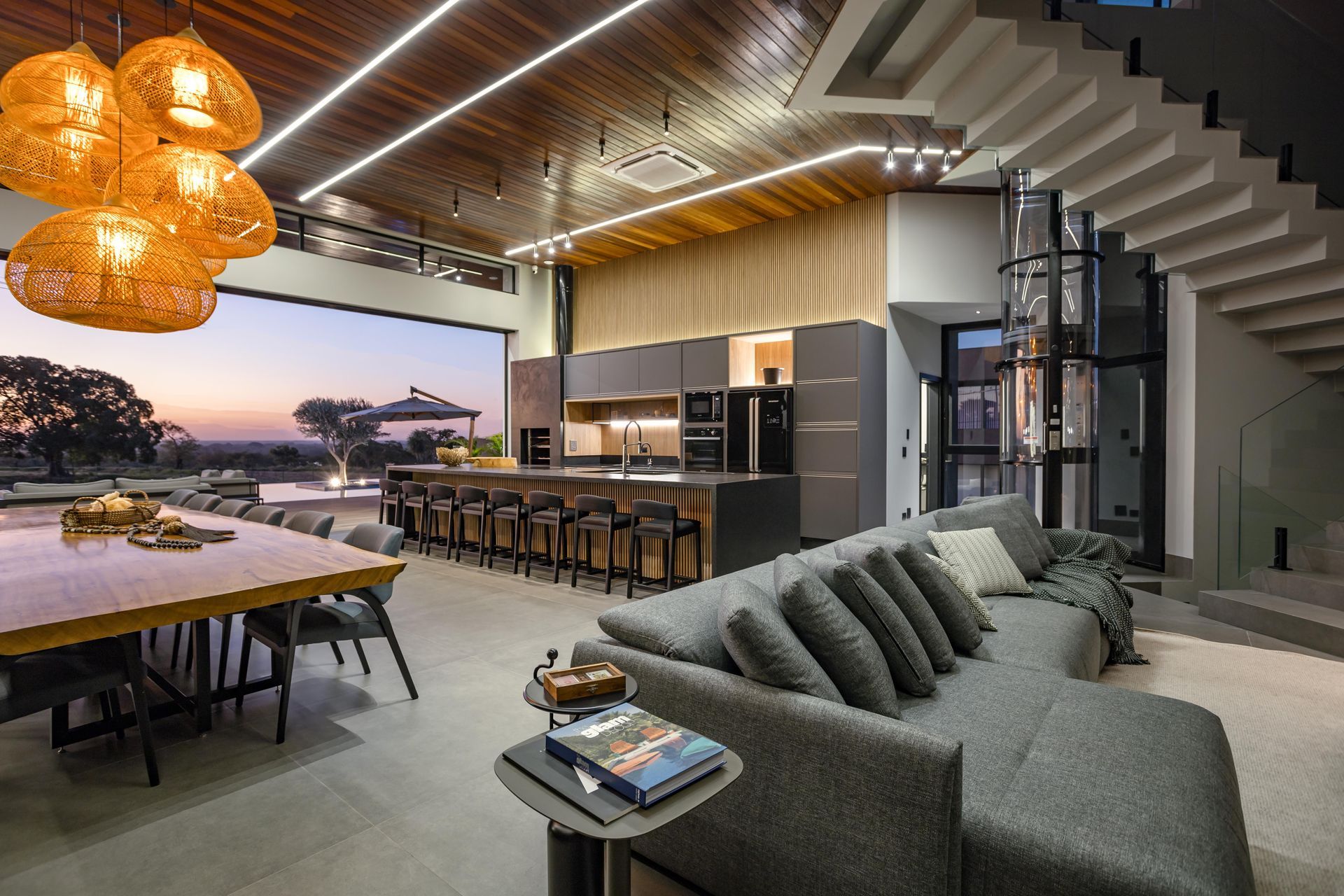 Luxuosa sala de estar em plano aberto com cozinha, sala de jantar e sala de estar. Possui janelas amplas, móveis modernos e uma escada em espiral.