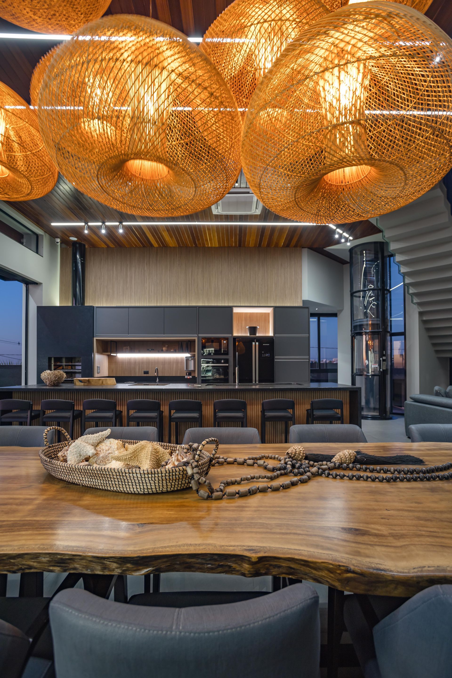 Sala de jantar com mesa de madeira, luminárias de vime e cozinha ao fundo.