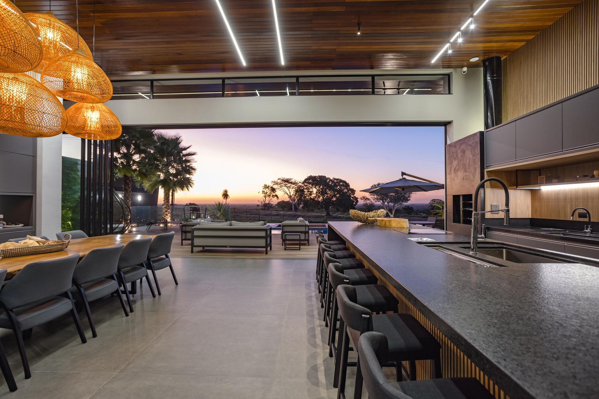 Cozinha moderna com vista aberta para o pôr do sol e área de estar ao ar livre. Possui uma ilha longa com bancos altos e uma grande mesa de jantar.
