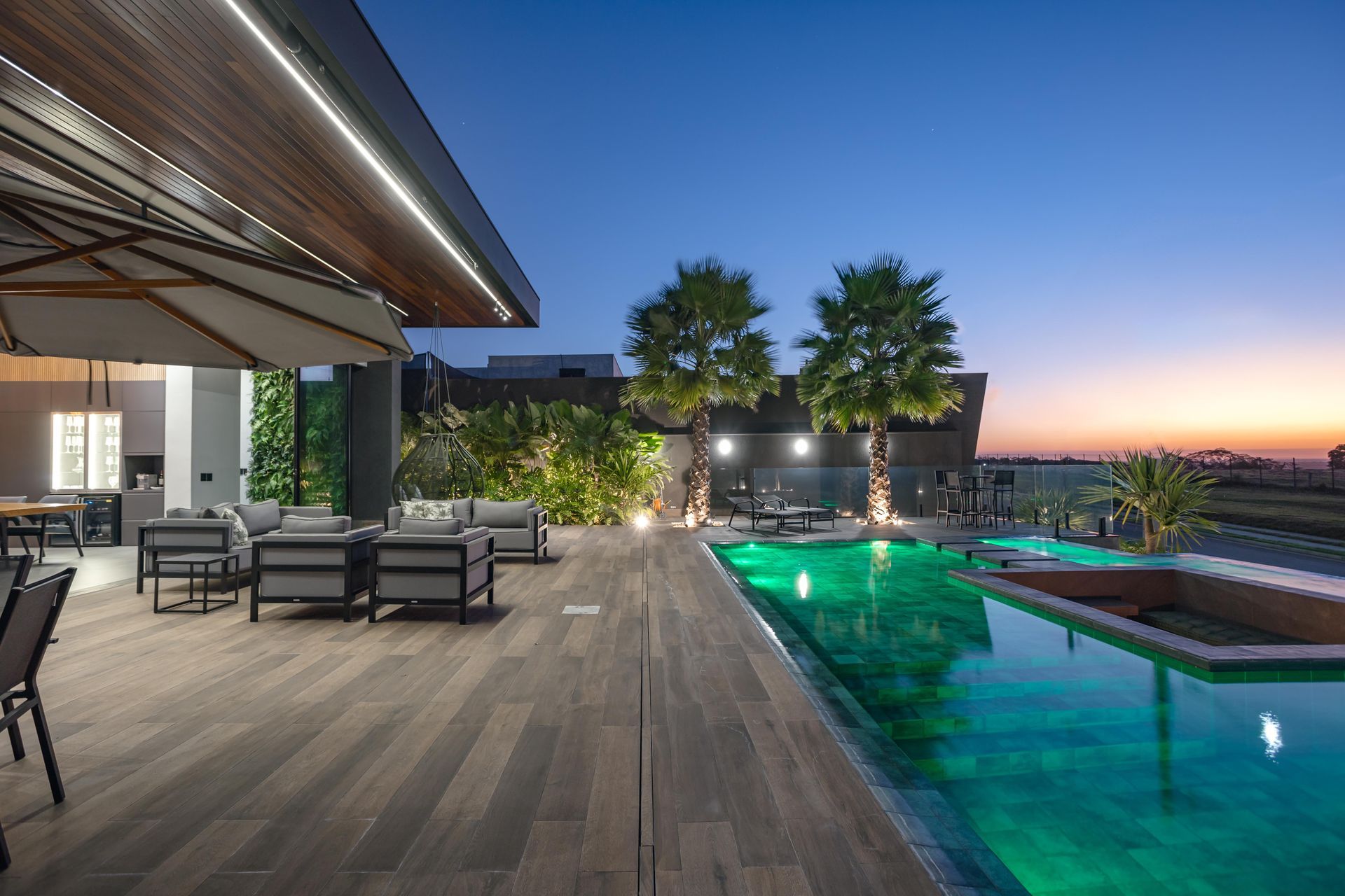 Piscina na cobertura com área de estar ao entardecer; palmeiras, água verde e deck de madeira.