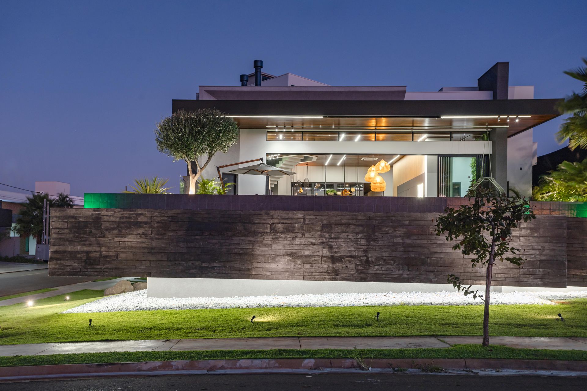 Casa moderna ao entardecer com fachada iluminada, parede de concreto e paisagismo.