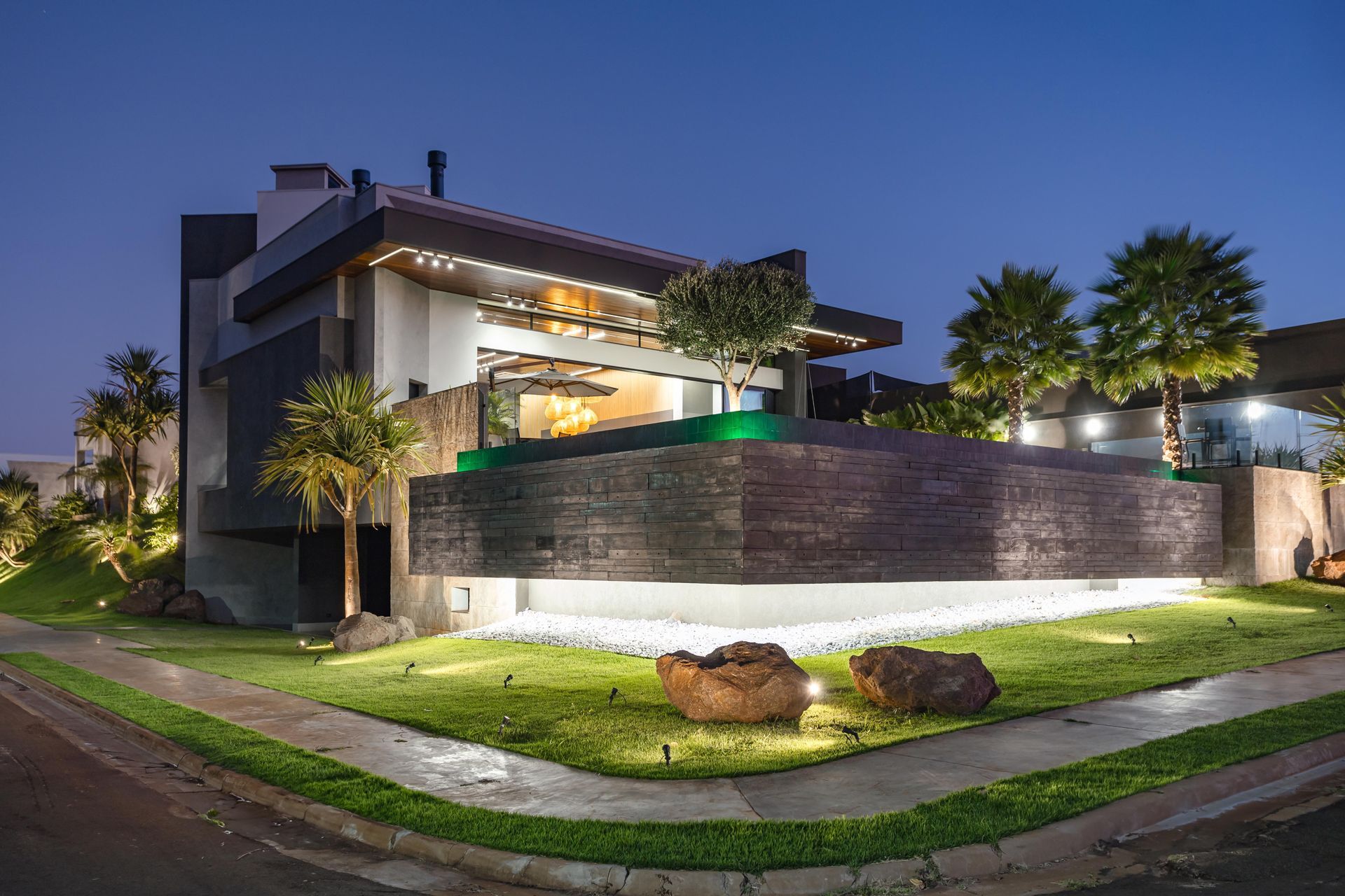 Exterior de uma casa moderna iluminado à noite com paisagismo e detalhes em pedra.