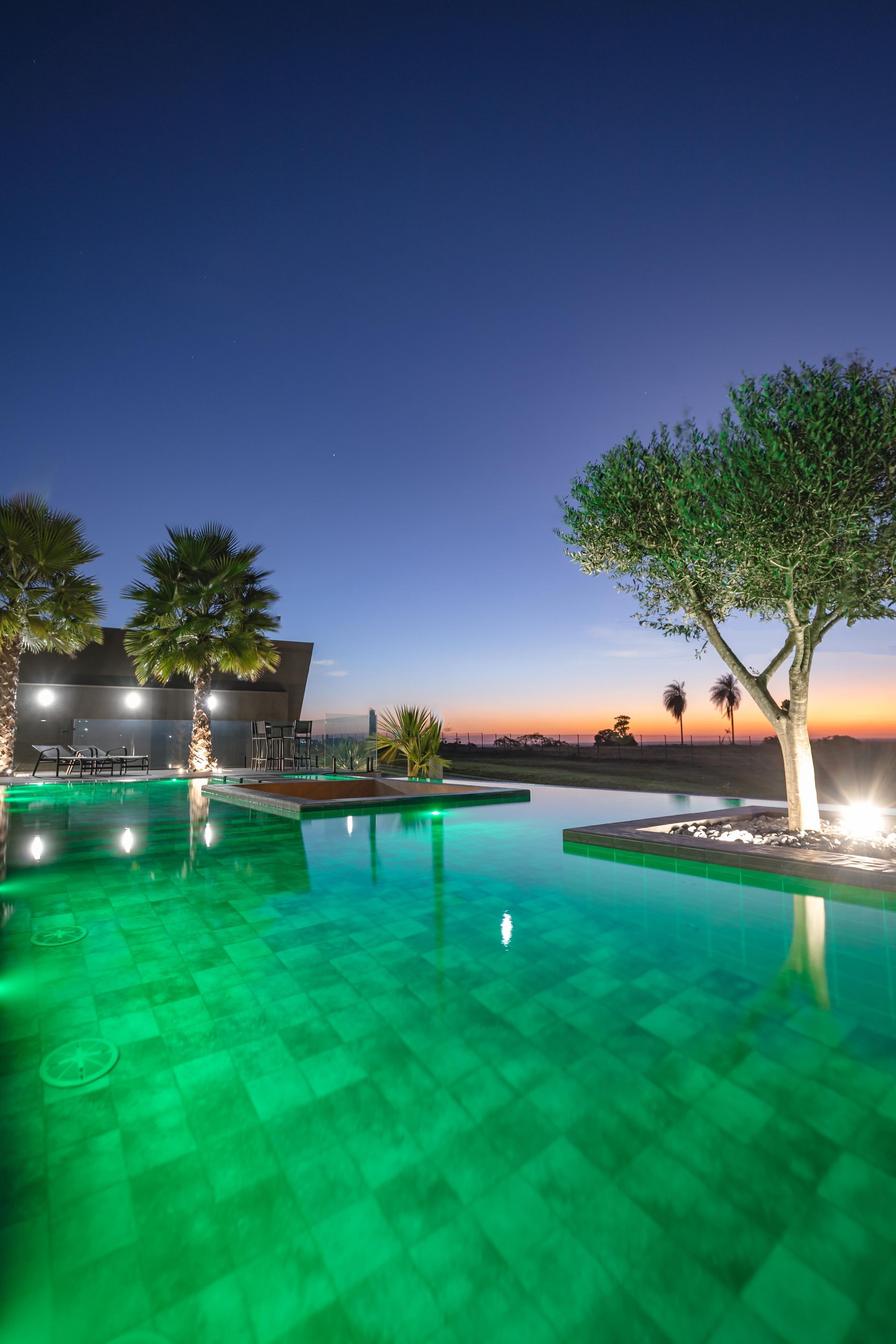 Piscina iluminada com luzes verdes, árvores recortadas contra o céu ao entardecer.