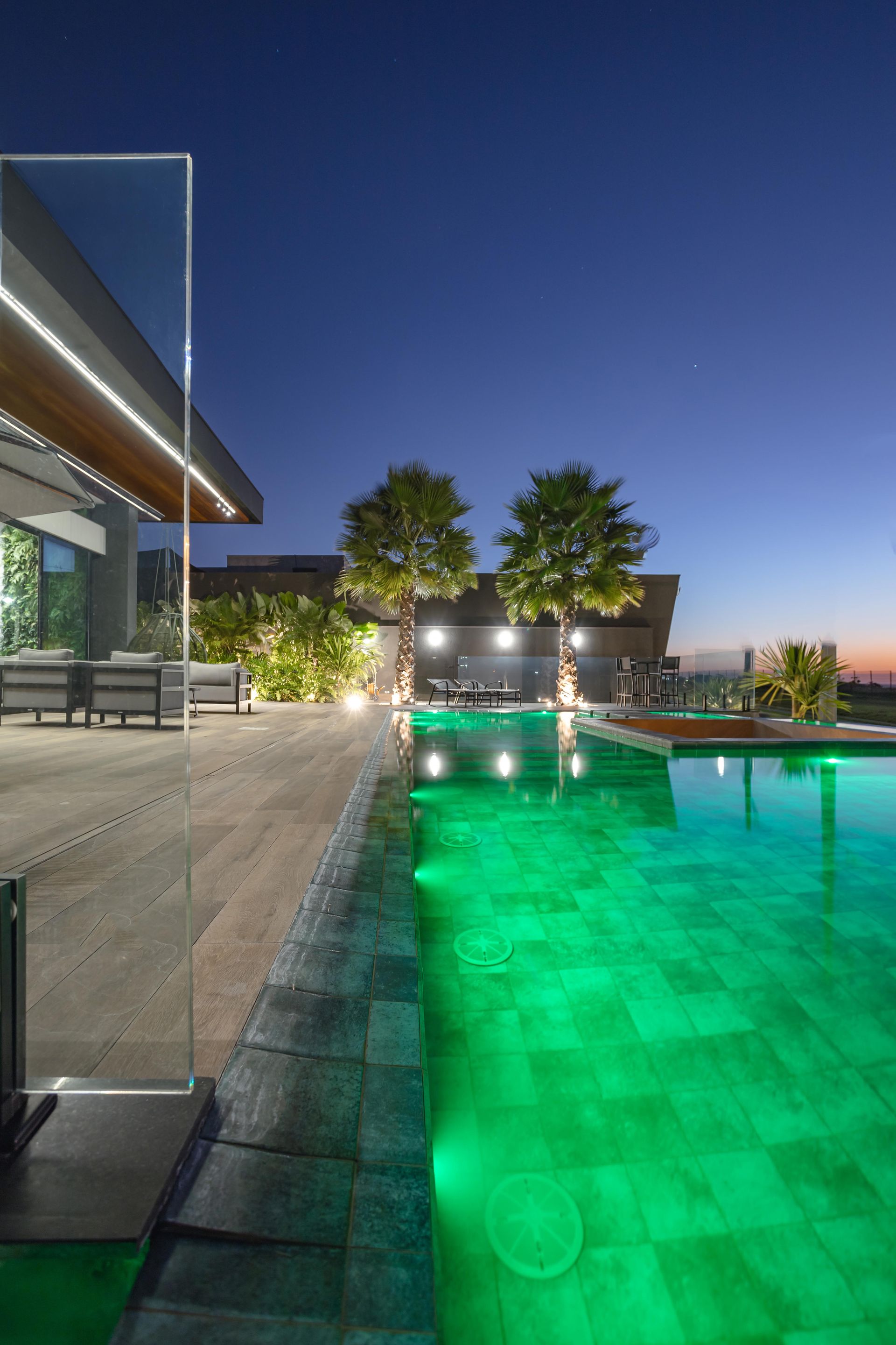 Cena noturna: piscina iluminada em verde, arquitetura moderna, árvores e céu crepuscular.