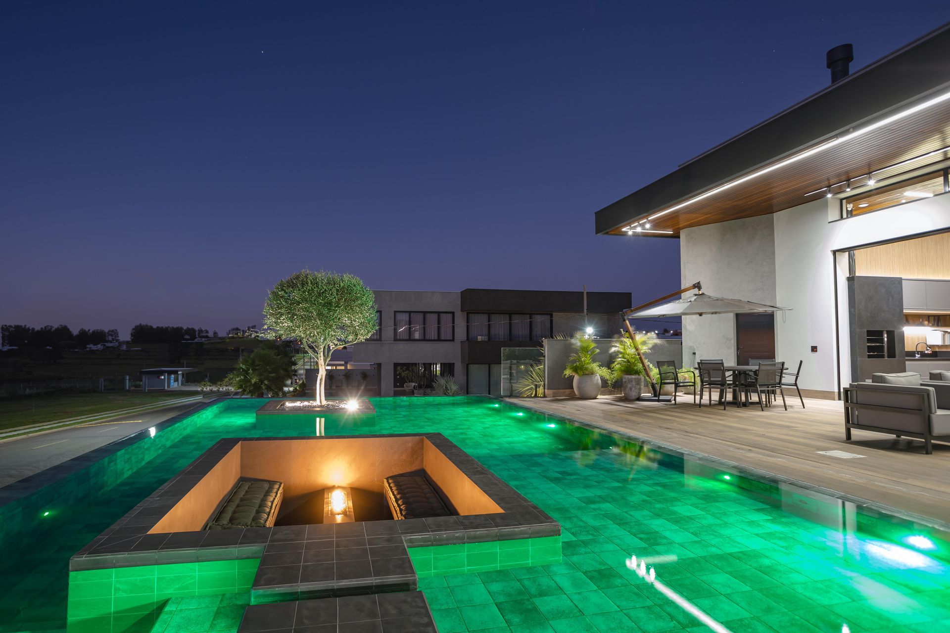 Vista noturna de uma casa luxuosa com piscina com iluminação verde e fogueira. Móveis de jardim visíveis.