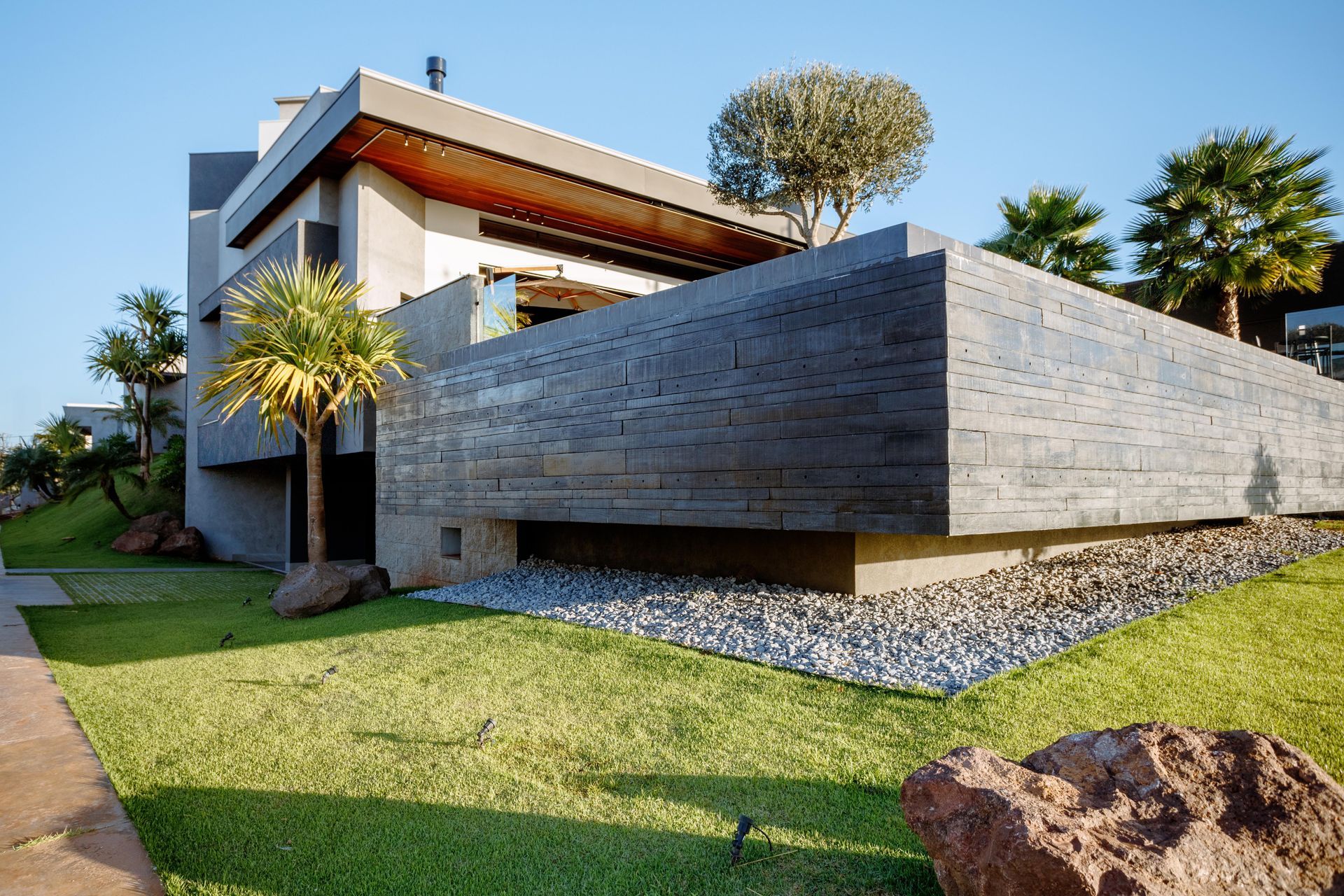 Casa cinza moderna com fachada de pedra, gramado verdejante e palmeiras em um dia ensolarado.