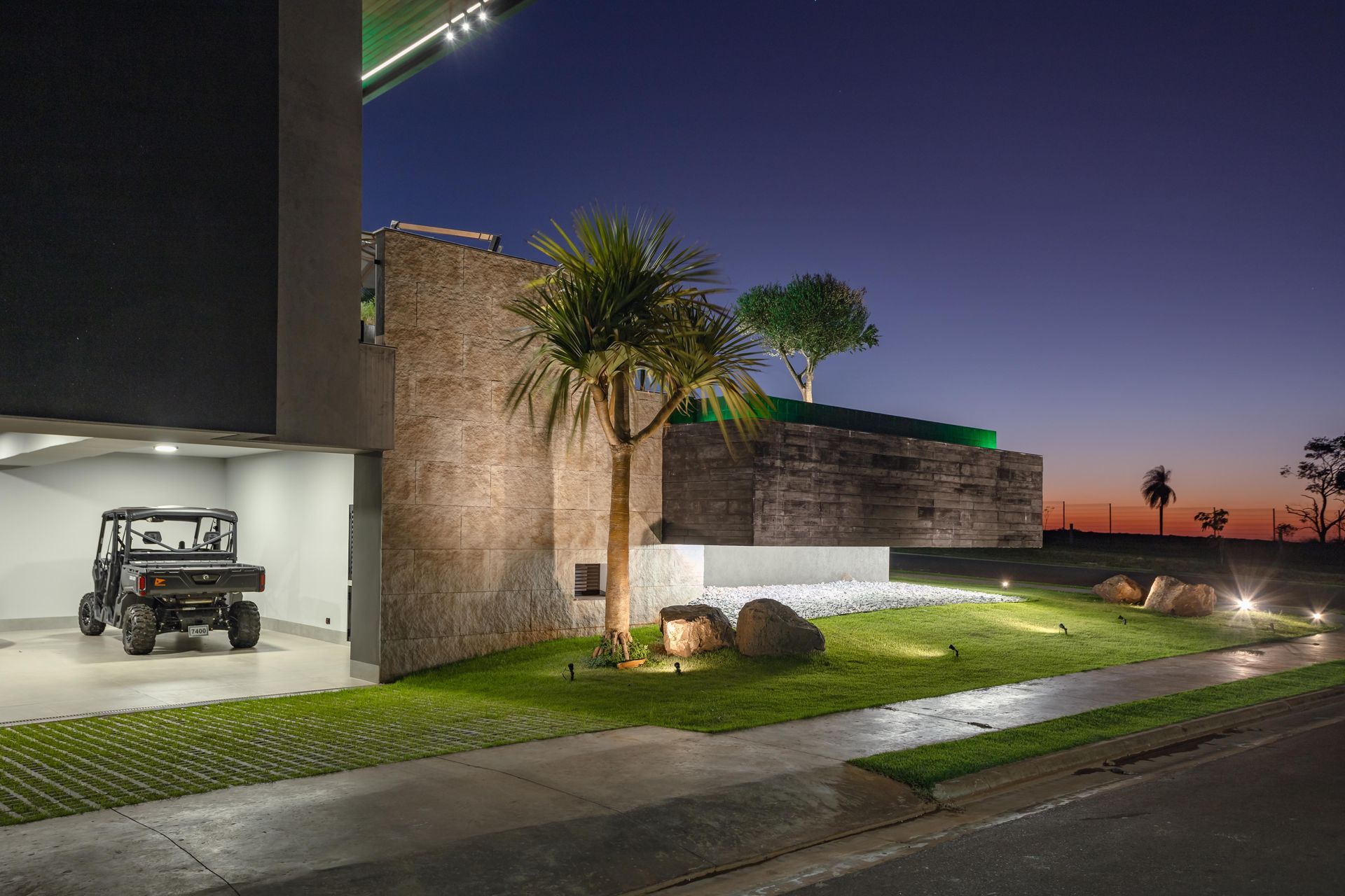 Exterior de casa moderna ao entardecer, com garagem e veículo utilitário. Gramado com árvores e detalhes em pedras.