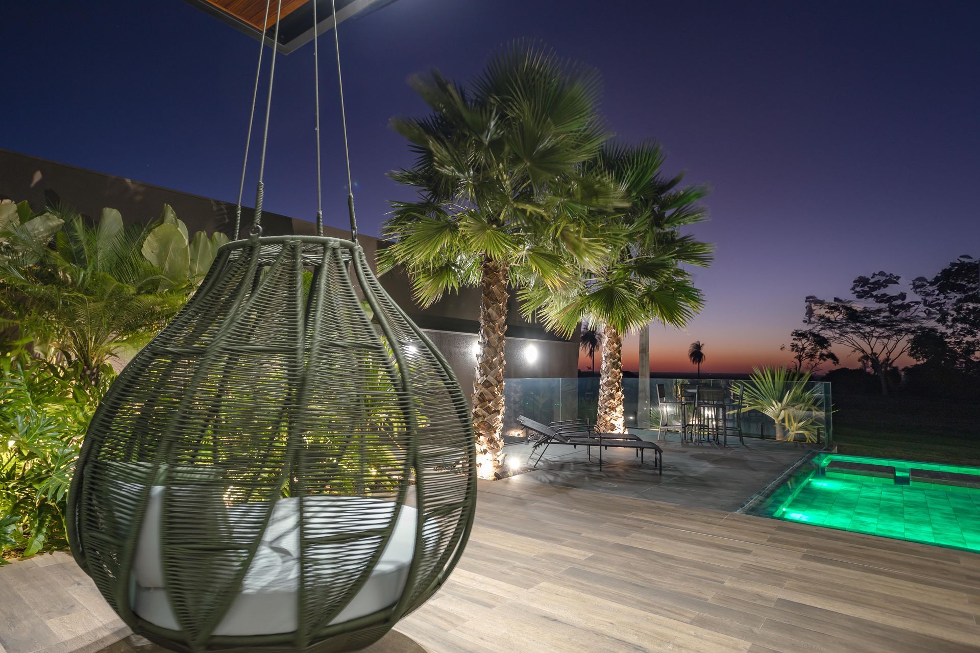Cena ao ar livre ao entardecer com uma cadeira suspensa, palmeiras e piscina iluminada, criando uma atmosfera relaxante.