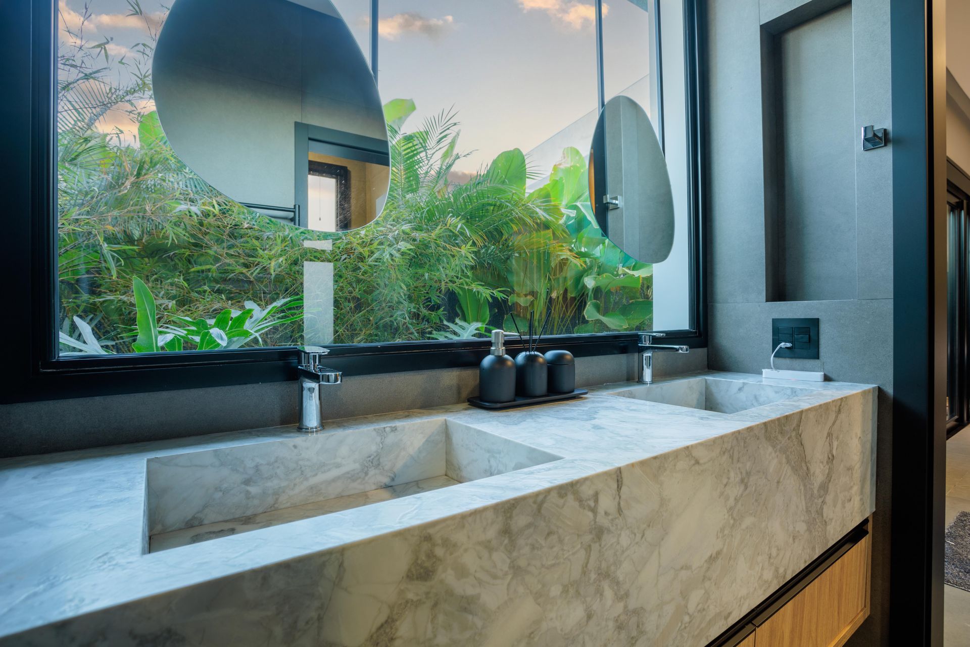 Banheiro com bancada de mármore, pias duplas e janela grande com vista para a área verde.