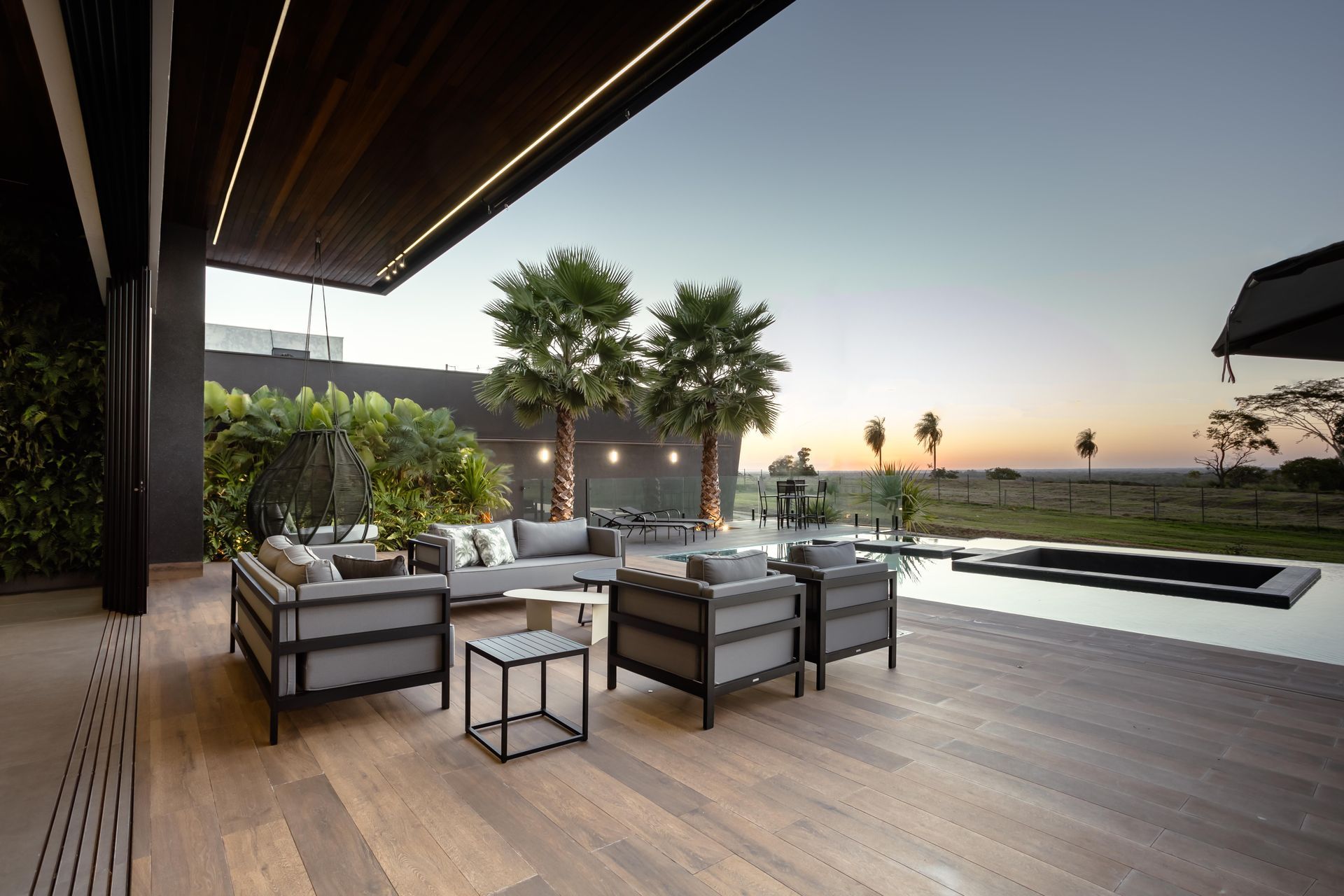 Pátio externo com móveis cinza, palmeiras e uma piscina escura ao pôr do sol.