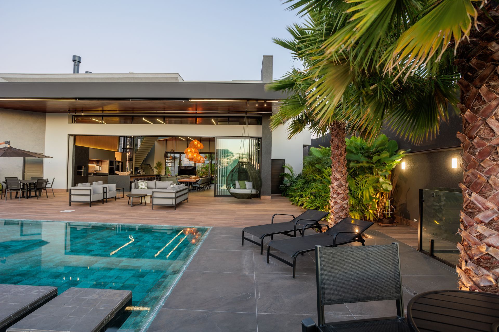 Pátio à beira da piscina com uma casa moderna, piscina, espreguiçadeiras, palmeiras e iluminação noturna.