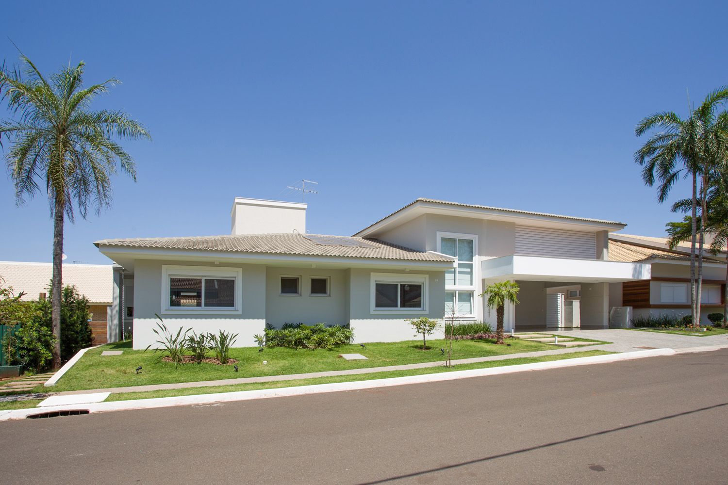 Casa cinza claro com palmeiras e entrada de automóveis sob um céu azul claro.