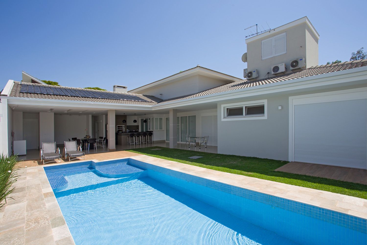 Área de piscina com uma casa, piscina azul, grama verde e céu ensolarado.