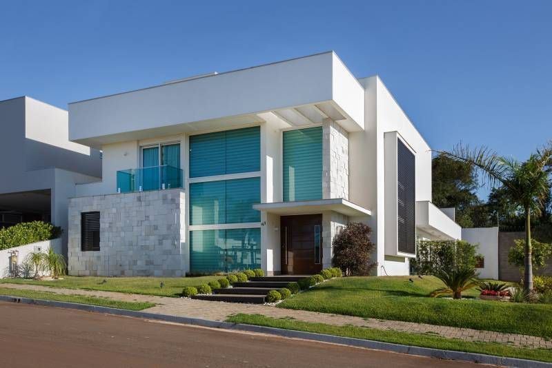 Casa branca moderna com janelas azuis, porta de madeira e pequenos degraus que levam à entrada, situada em um quintal verde.