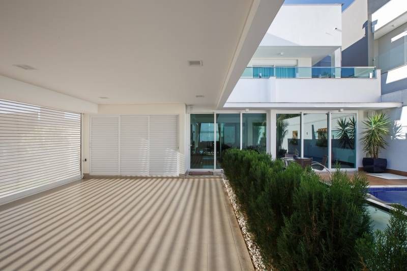 Pátio coberto com paredes brancas e uma fileira de arbustos verdes em frente a um espaço com paredes de vidro.