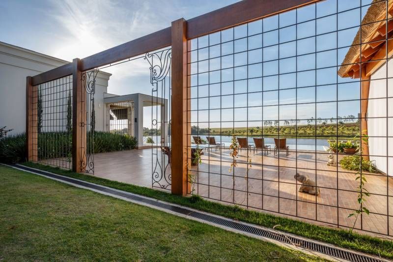 Cerca de tela de arame com estrutura de madeira com vista para um pátio, lago e cadeiras sob um céu ensolarado.