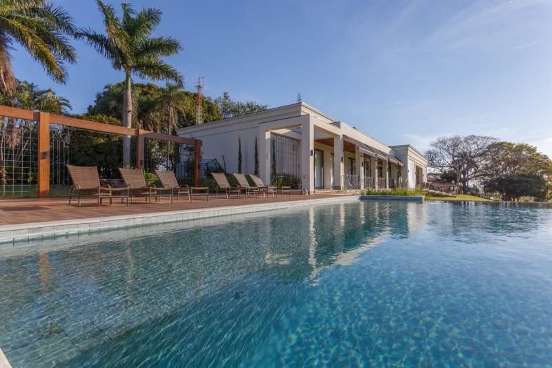 Piscina em frente a um prédio branco com espreguiçadeiras e palmeiras sob um céu azul brilhante.