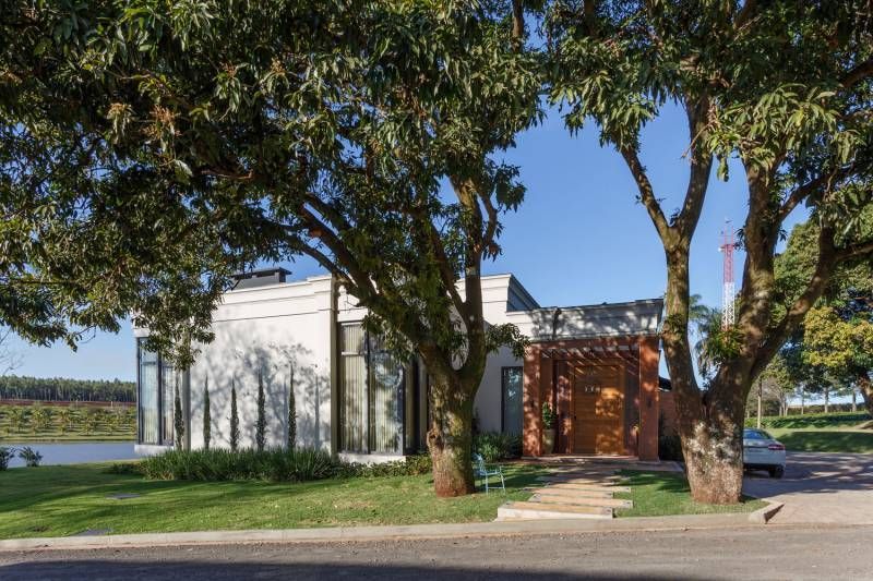 Casa moderna branca parcialmente escondida por árvores, céu azul e grama verde.