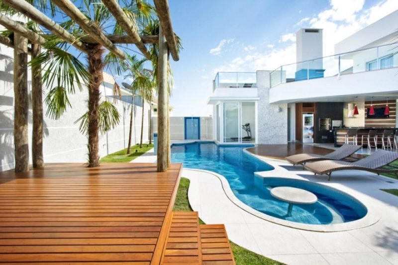 Deck de madeira e piscina com vista para uma moderna casa branca com palmeiras.