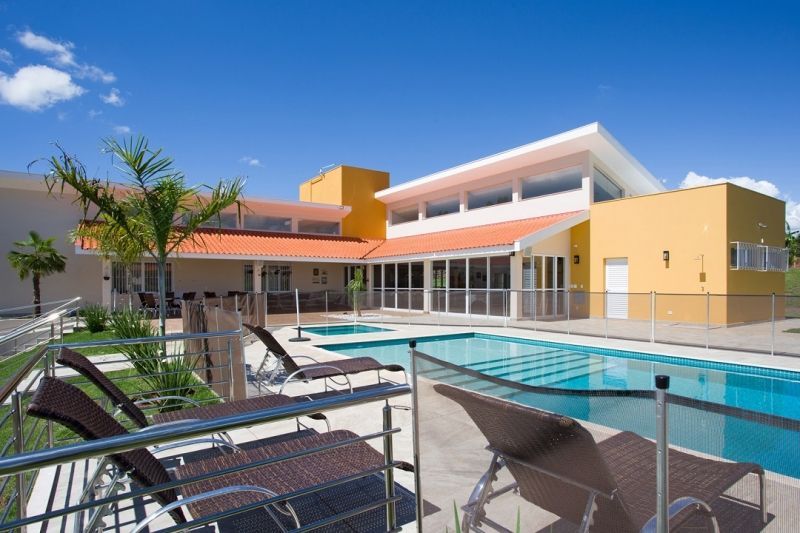 Área da piscina com espreguiçadeiras, prédio com paredes amarelas, céu azul.