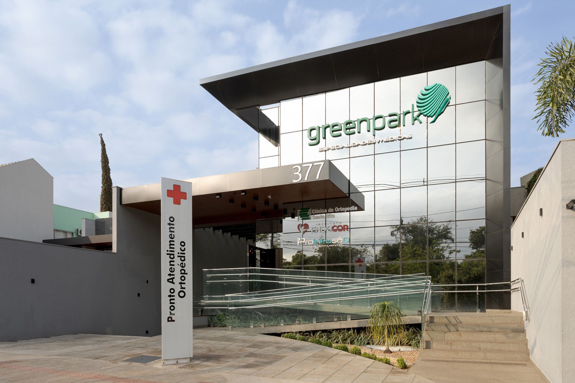 Prédio branco com detalhes em vermelho, placa verde e persianas verticais.