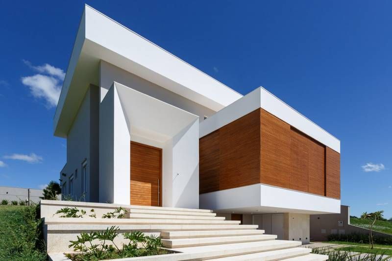 Casa branca moderna com detalhes em madeira, degraus que levam à entrada contra um céu azul.